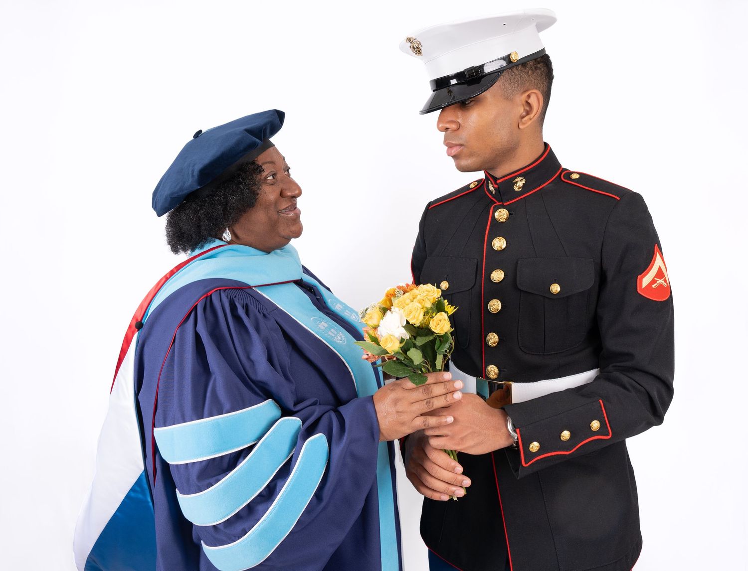 A mother and son posing on a white backdrop. The mother is in a graduation gown and the son is in his dress blues.