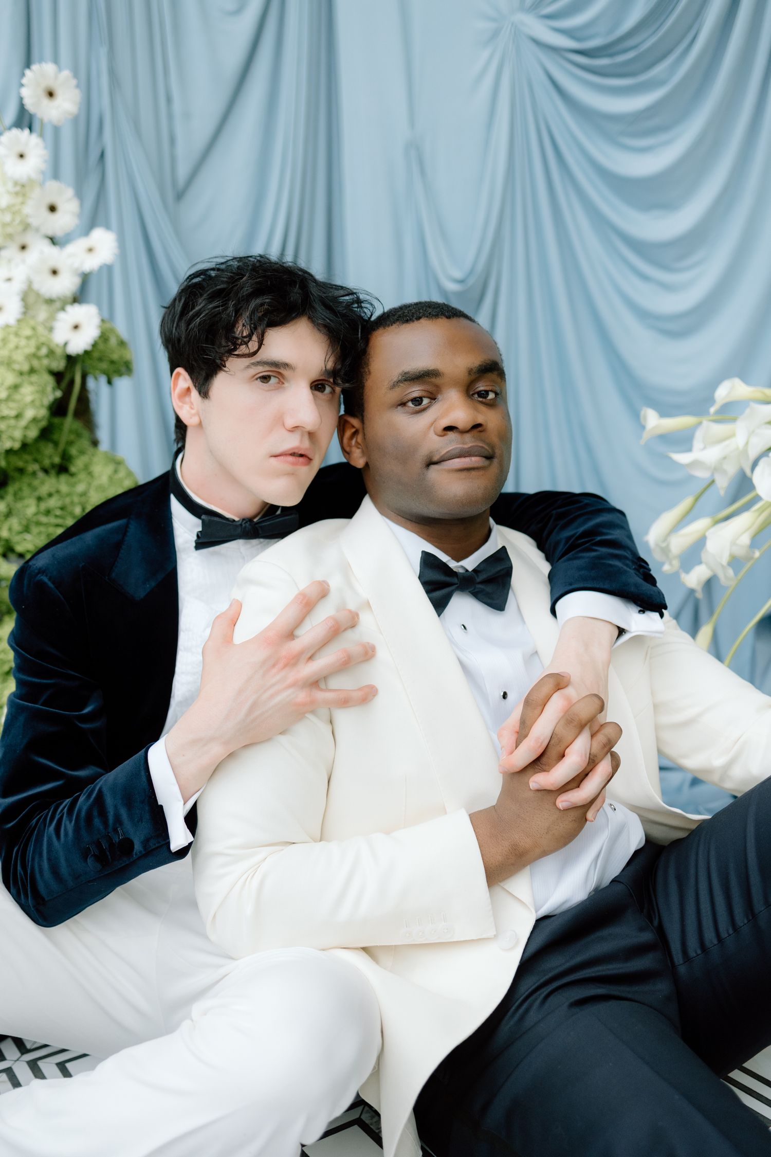 Two grooms in tuxedos embrace tenderly against a blue draped backdrop with white flowers at their wedding.
