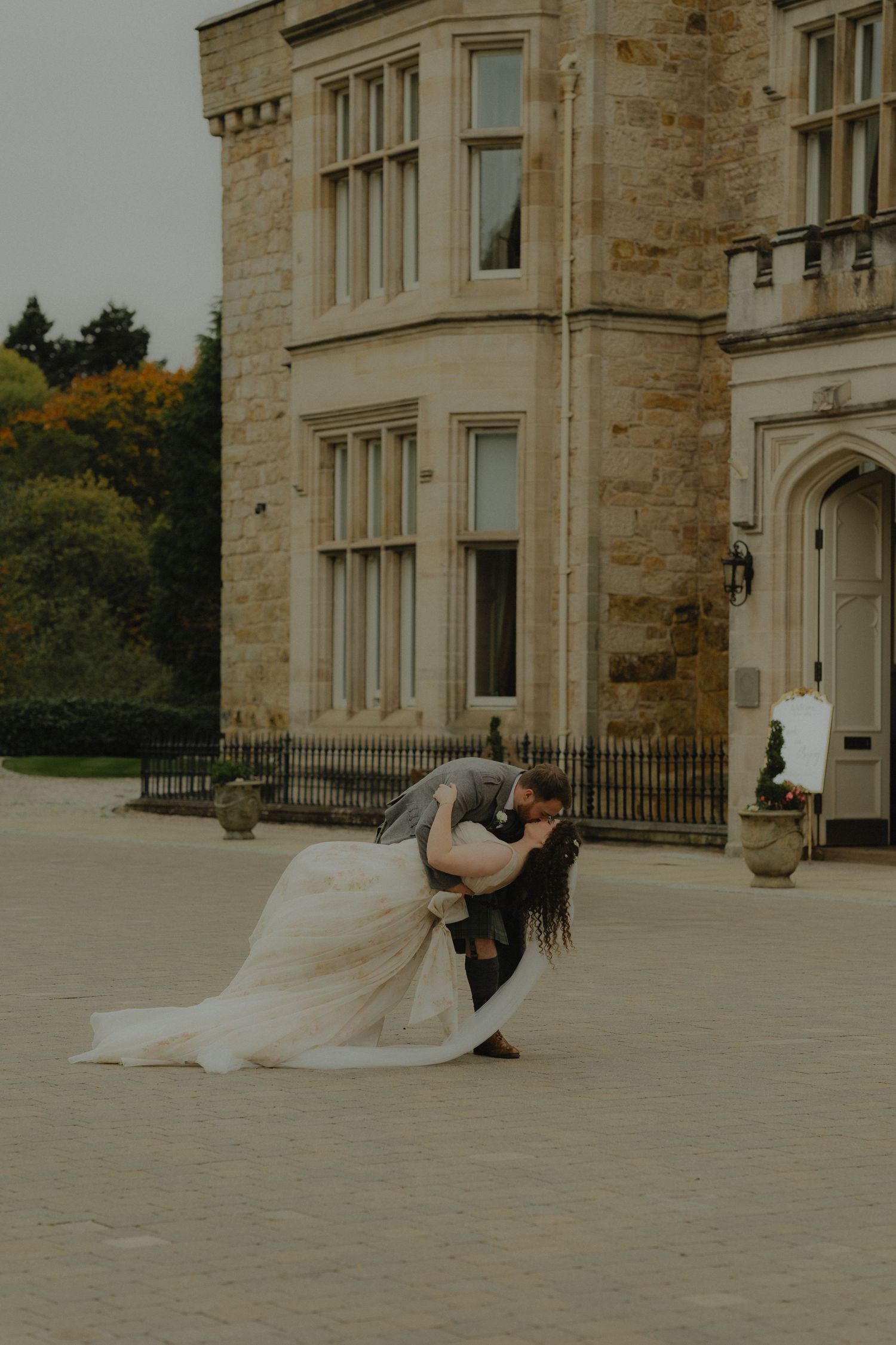 Groom dips bride in a romantic kiss outside a grand stone castle wedding venue.