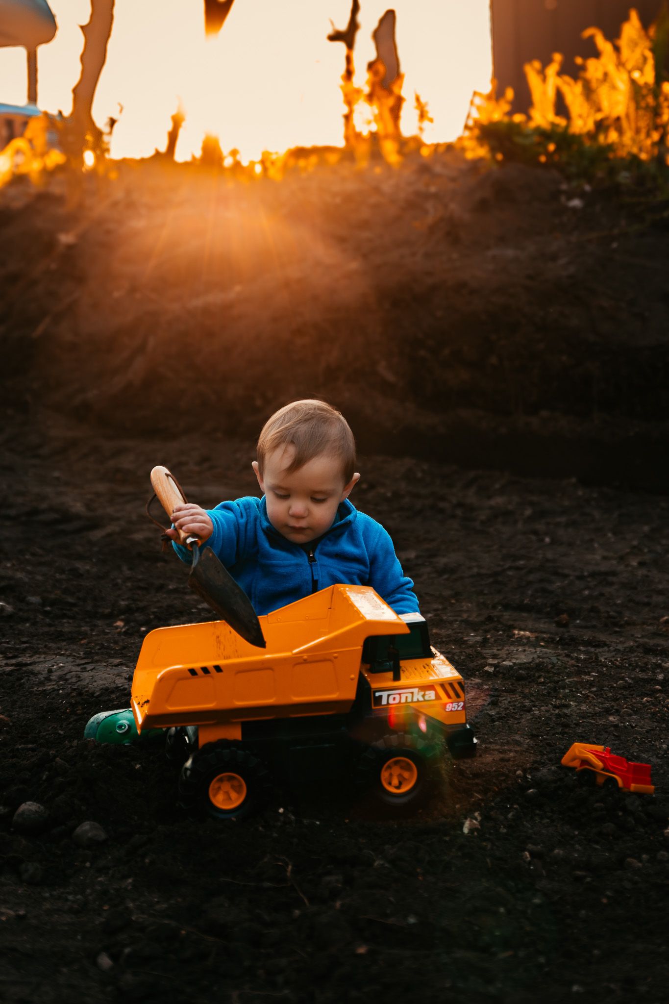 Toddler in blue shirt playing with orange Tonka dump truck in dirt at golden hour sunset.