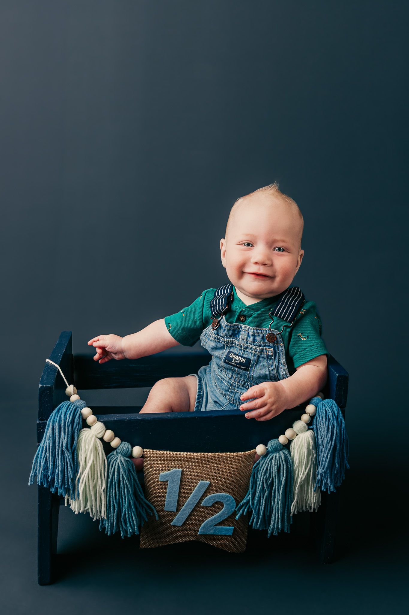Smiling baby in denim overalls sitting in a small blue wooden crib with a 1/2 birthday banner and tassels.
