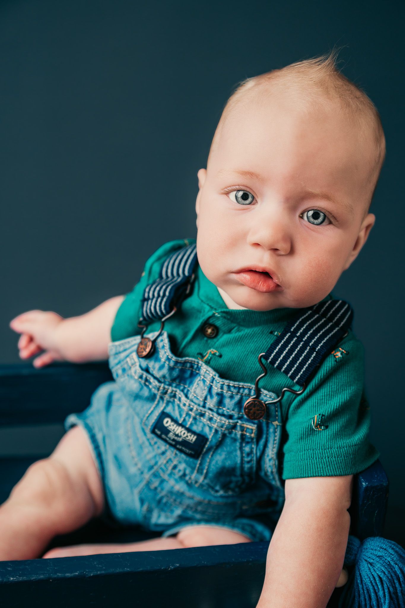 Adorable baby boy in denim OshKosh overalls and green polo shirt sitting and looking at camera.