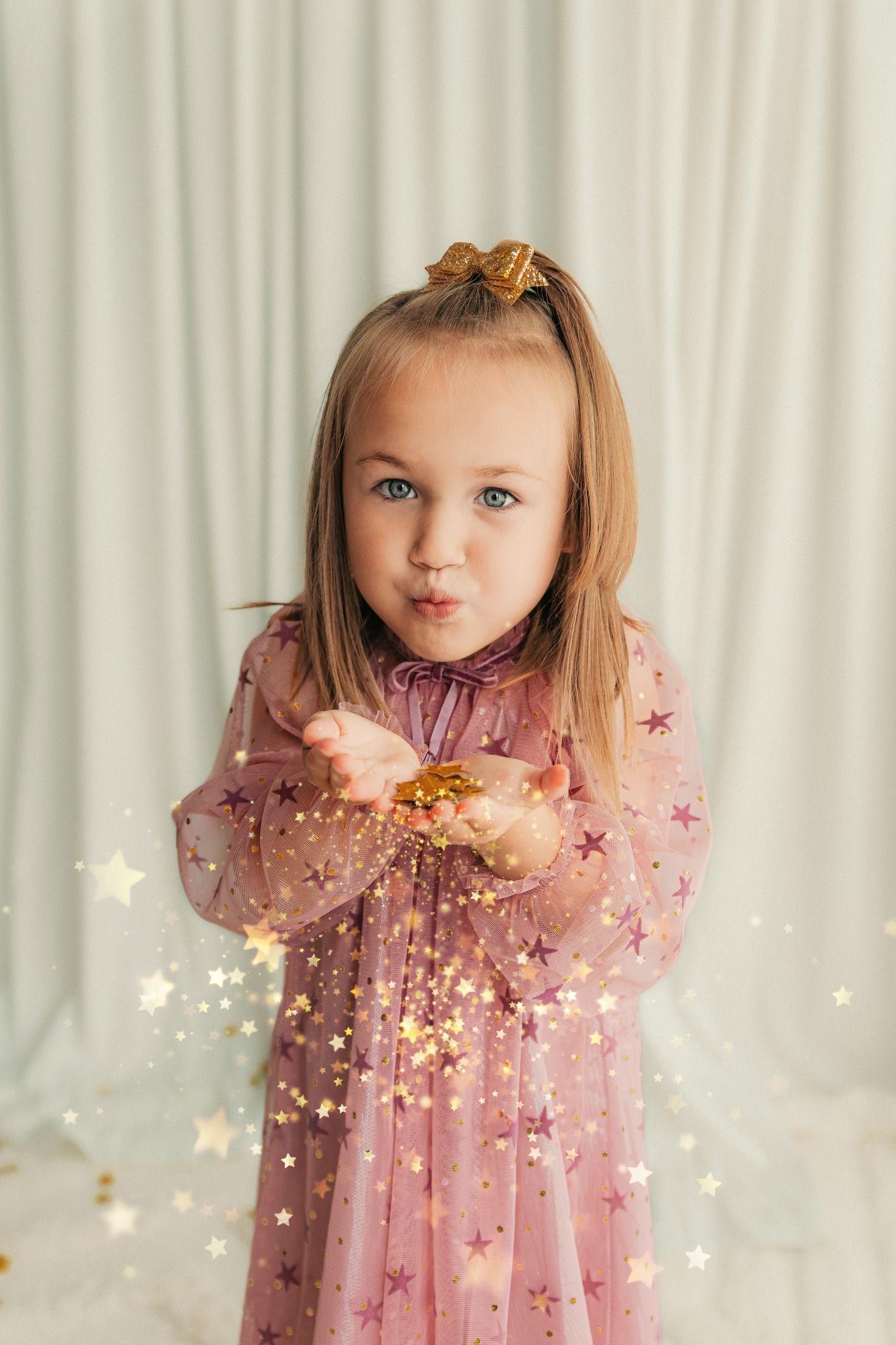 A young girl in a pink star-patterned dress blows golden glitter from her hands, wearing a gold bow hair clip.