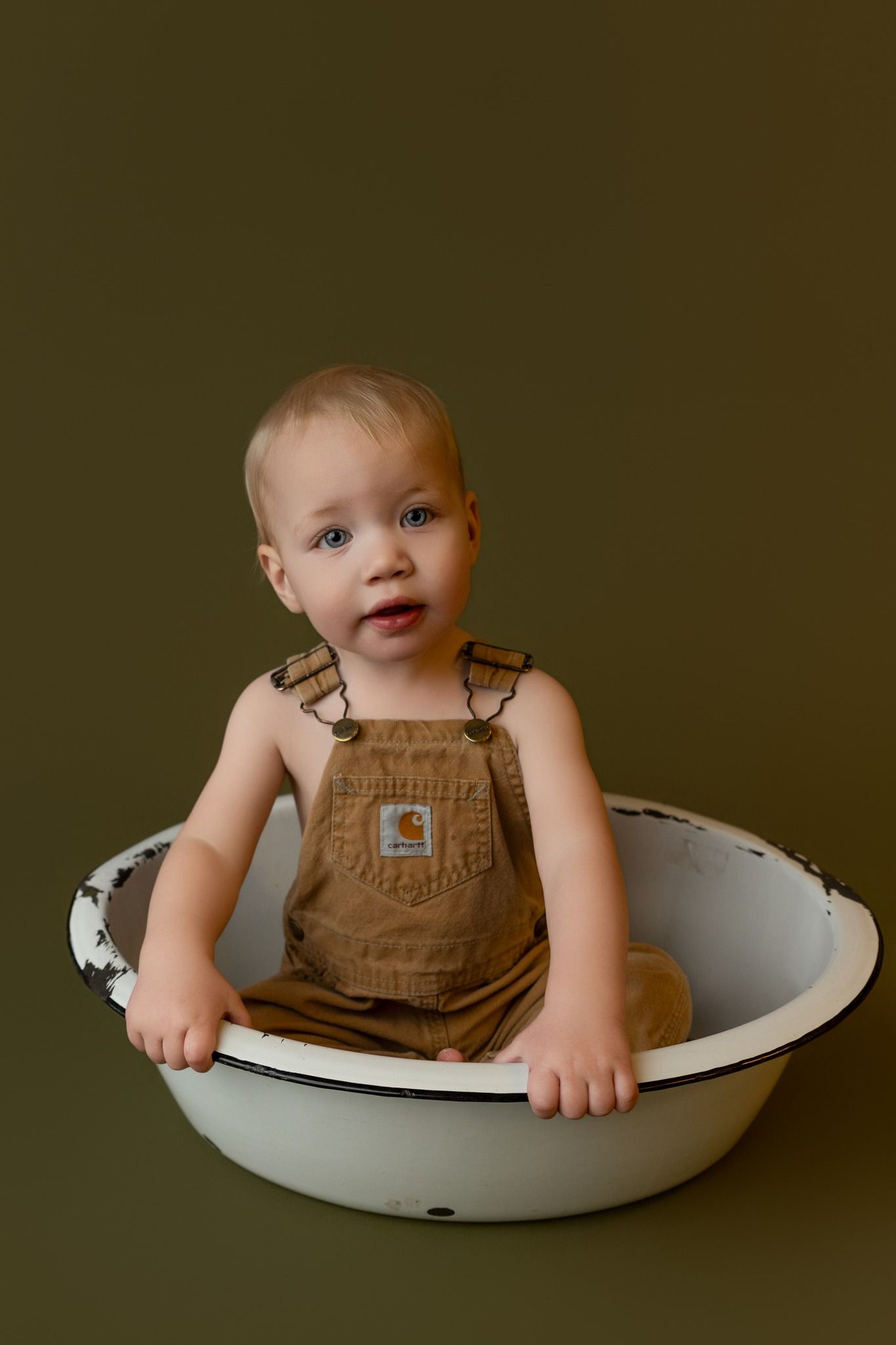 Baby in Carhartt overalls sitting in a vintage white enamel wash basin against an olive green background.