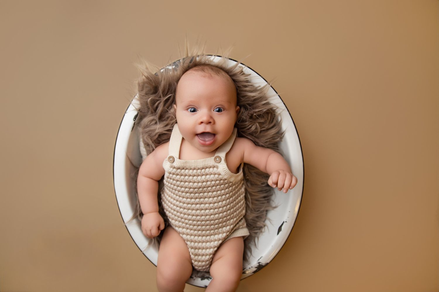 Newborn baby in knit romper lying on fluffy fur inside a white bowl against a tan background.