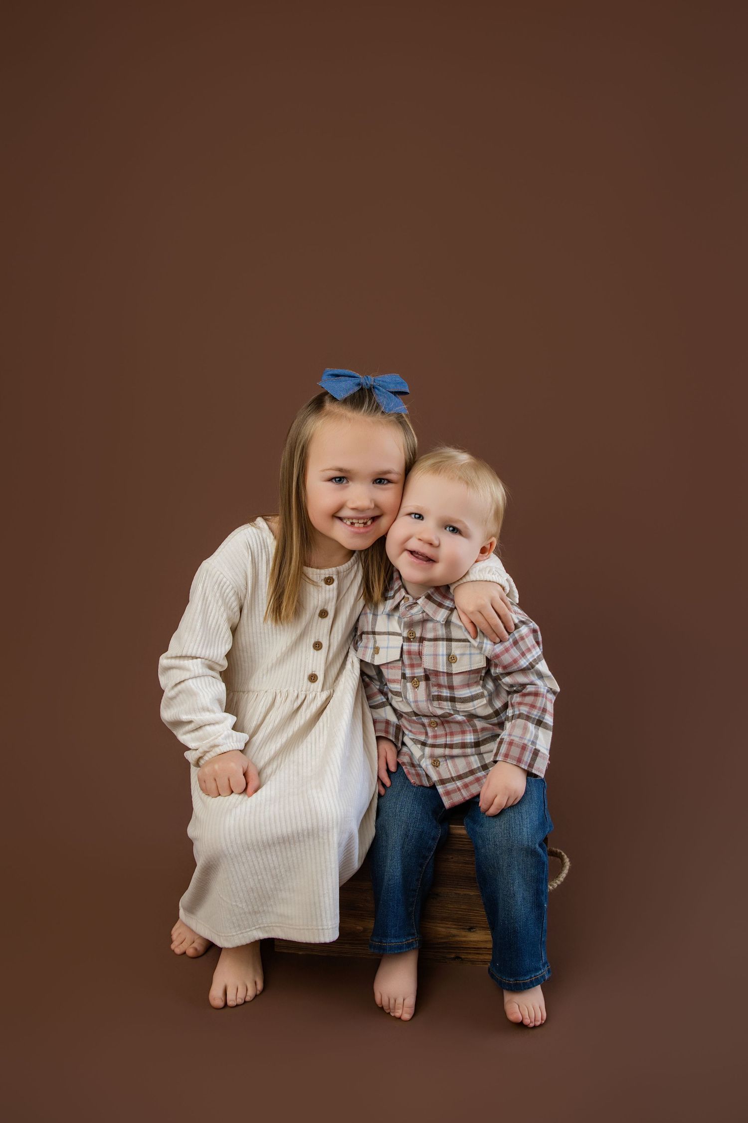 A smiling girl in a white dress with a blue bow hugs a toddler boy in plaid shirt and jeans on a brown backdrop.