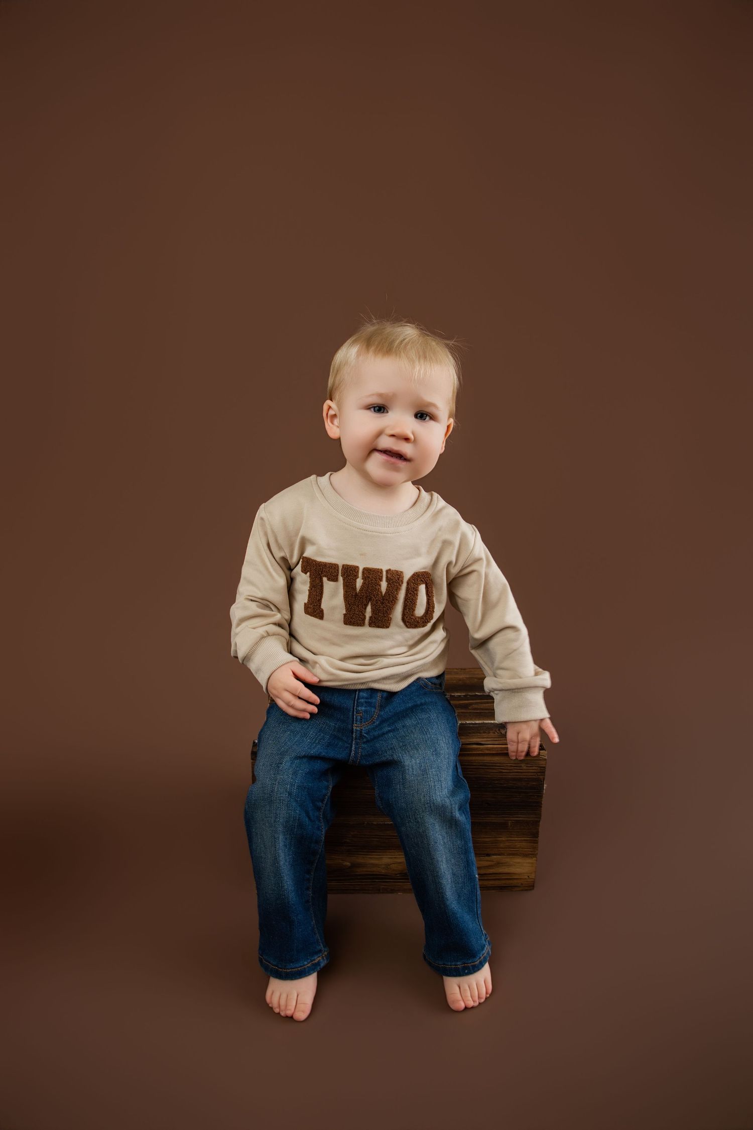 A smiling toddler wearing a 'TWO' sweatshirt and jeans sits on a wooden crate against a brown background.