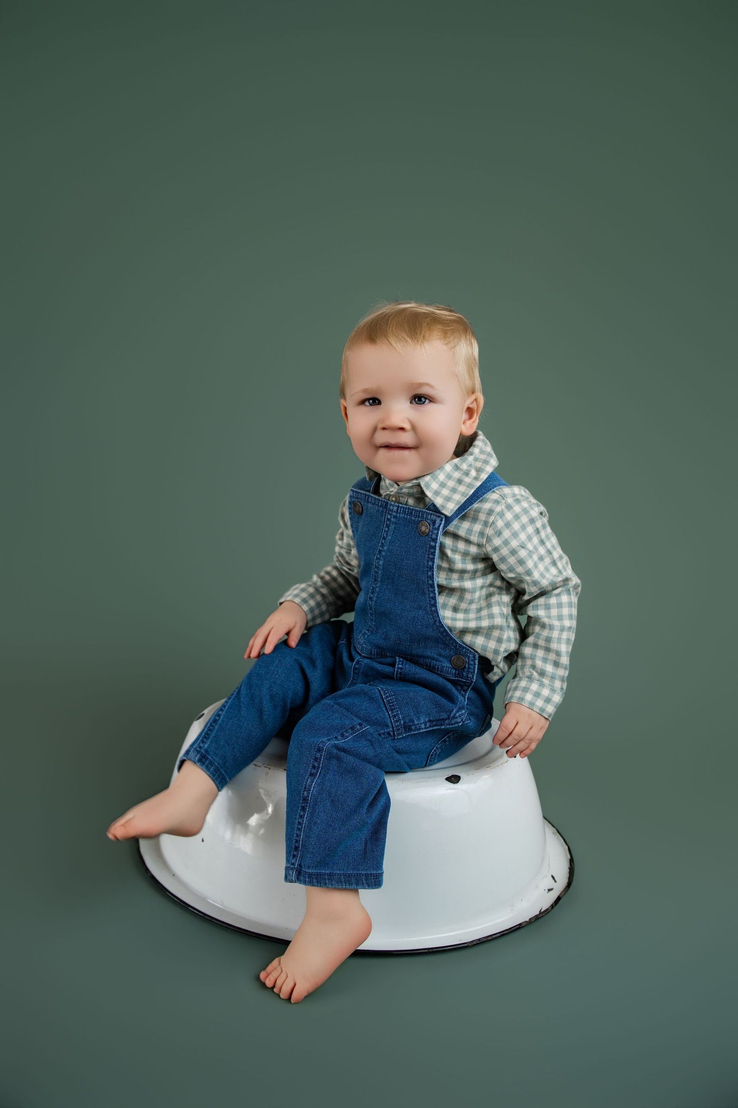 Toddler boy in denim overalls and plaid shirt sitting barefoot on white enamel basin against green background.