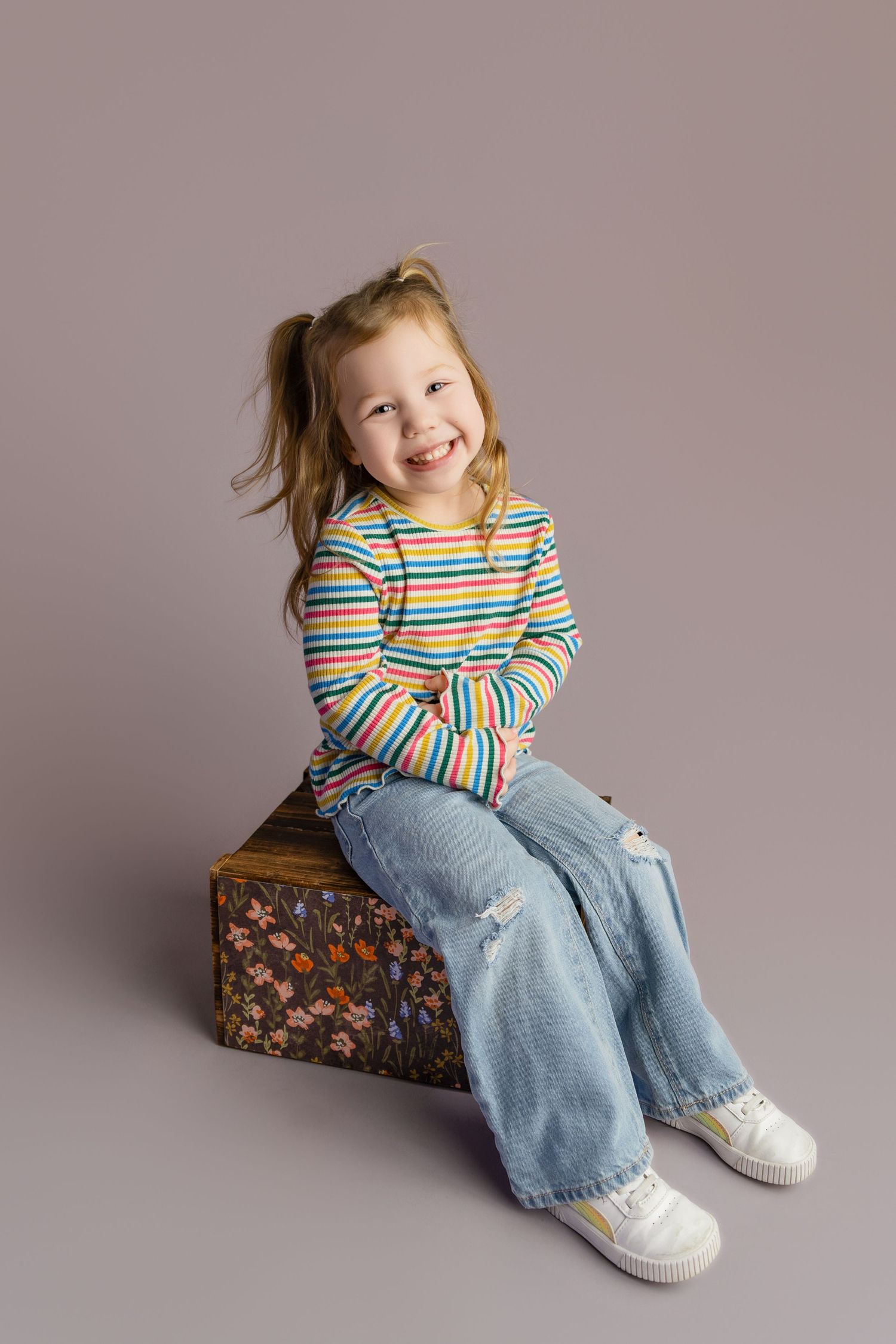 A cheerful young girl with pigtails sits on a floral box, wearing rainbow striped shirt and wide-leg jeans, smiling brightly.