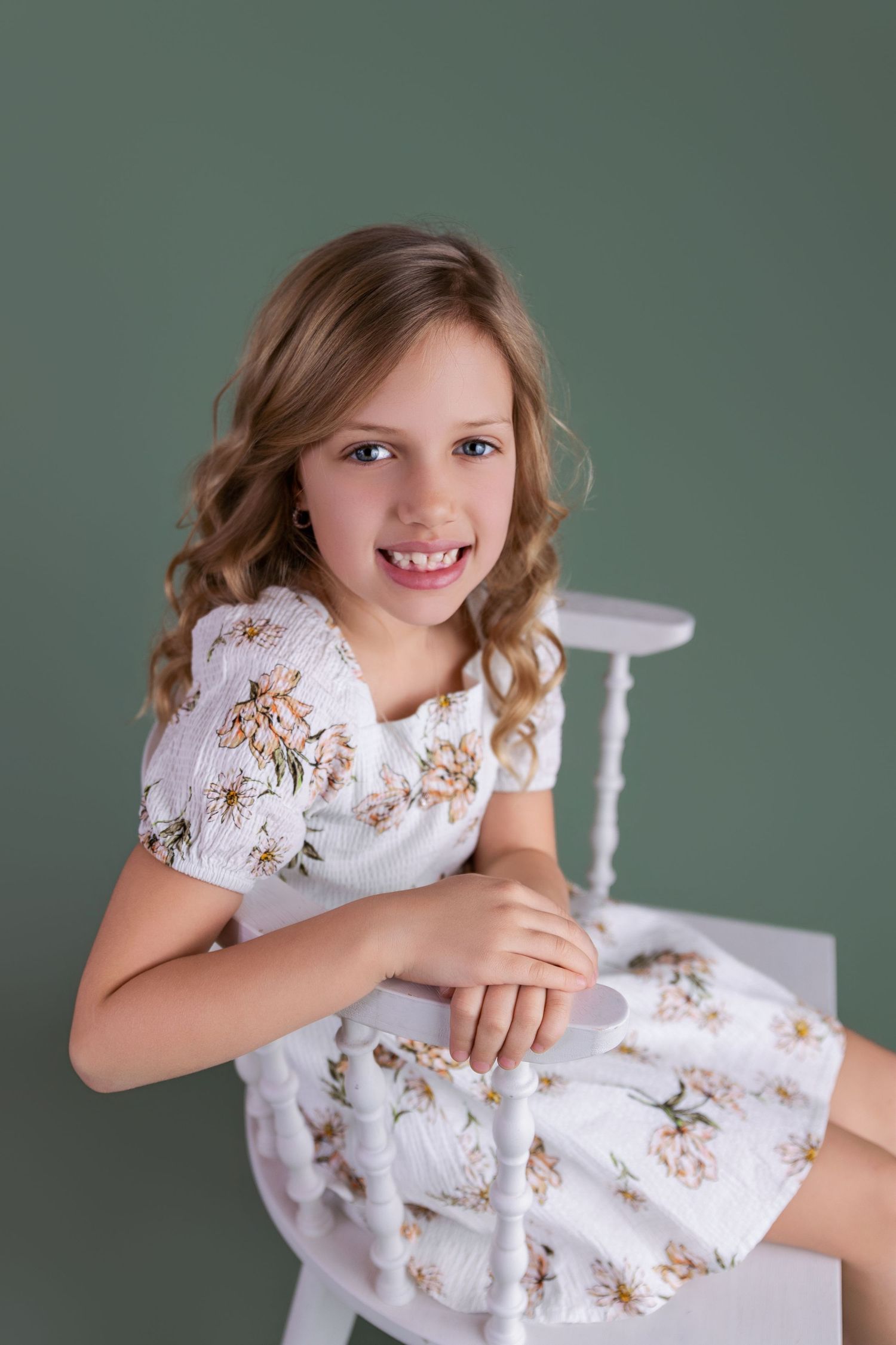 Smiling young girl with curly hair wearing a white floral dress, seated against a green background.