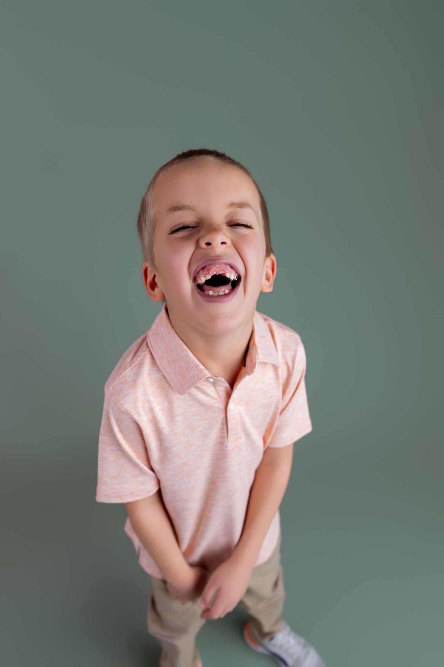 Young boy laughing with mouth open wide, wearing a pink polo shirt, standing against a gray-green background.