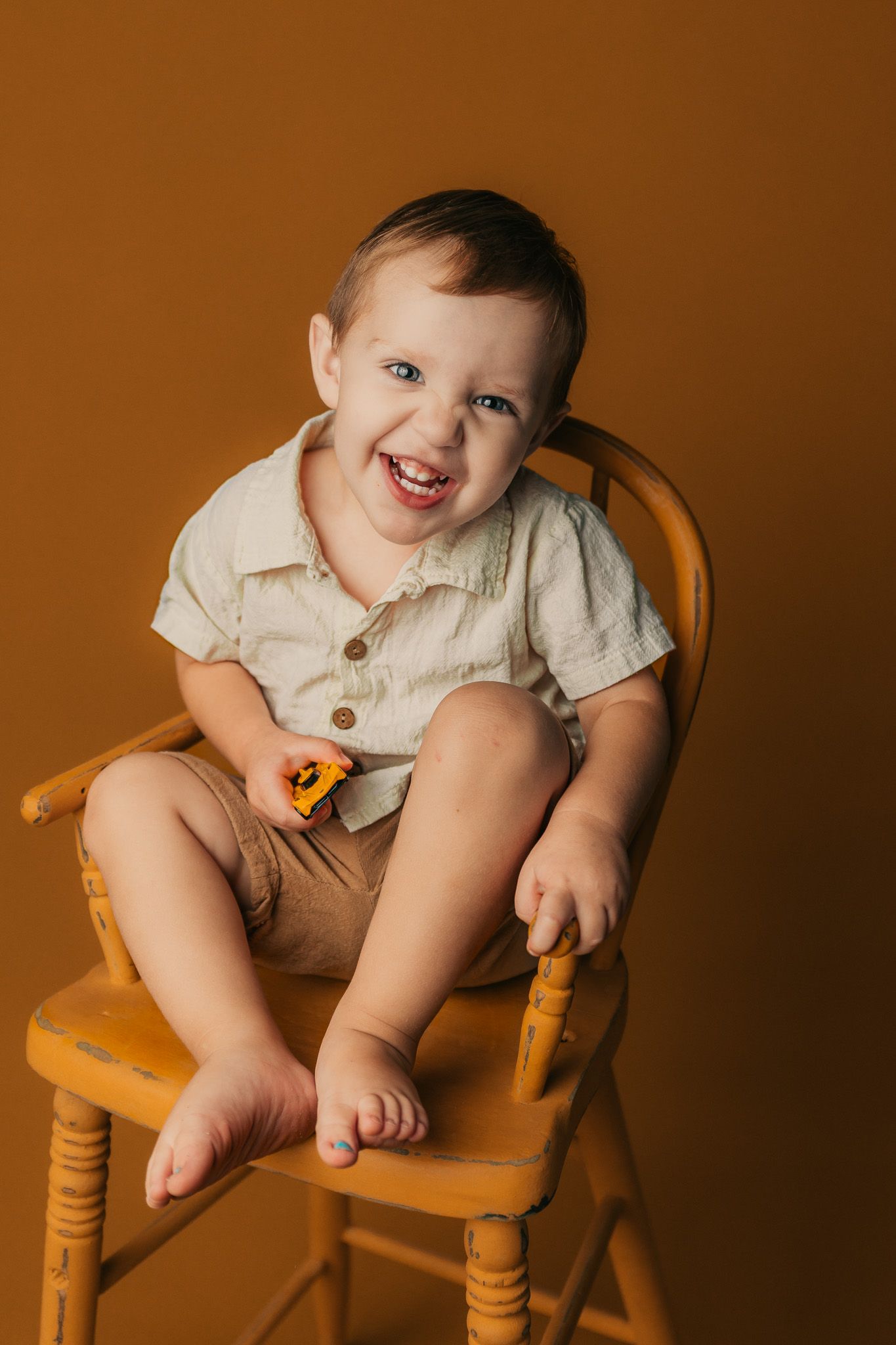 Smiling toddler boy with blue eyes sits on a yellow wooden high chair holding a toy car against a brown background.