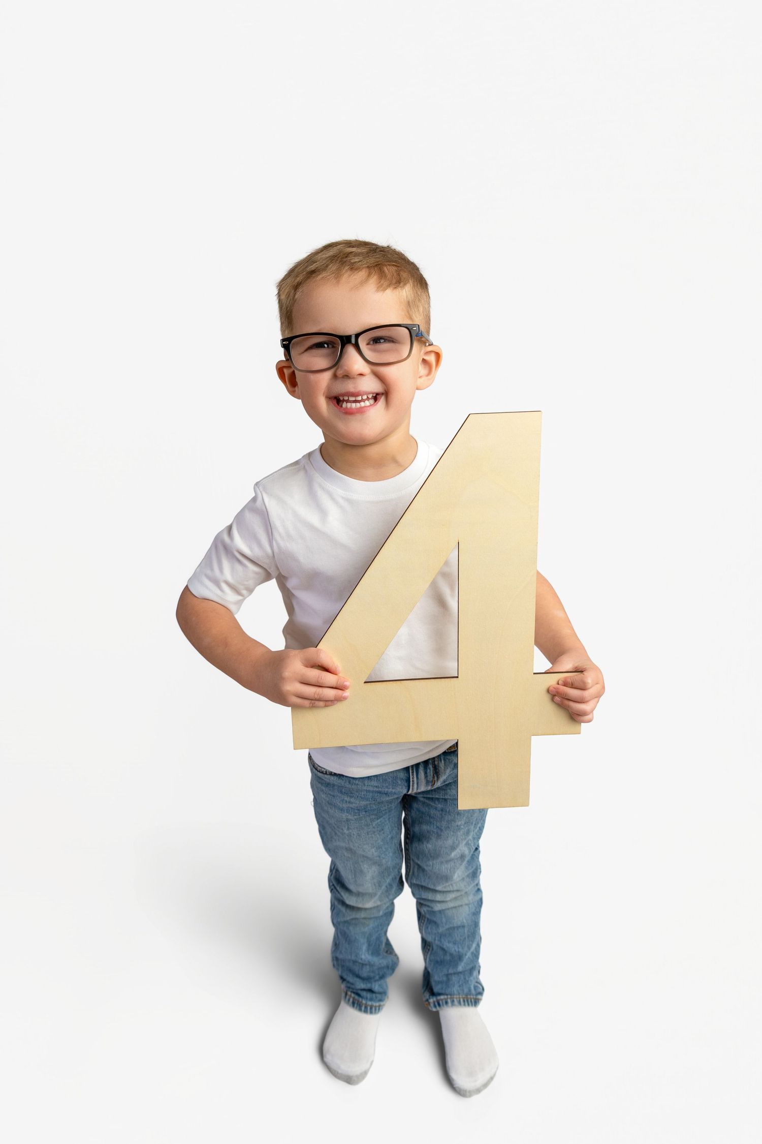 Smiling young boy wearing glasses and white t-shirt holding a large number 4 on white background.