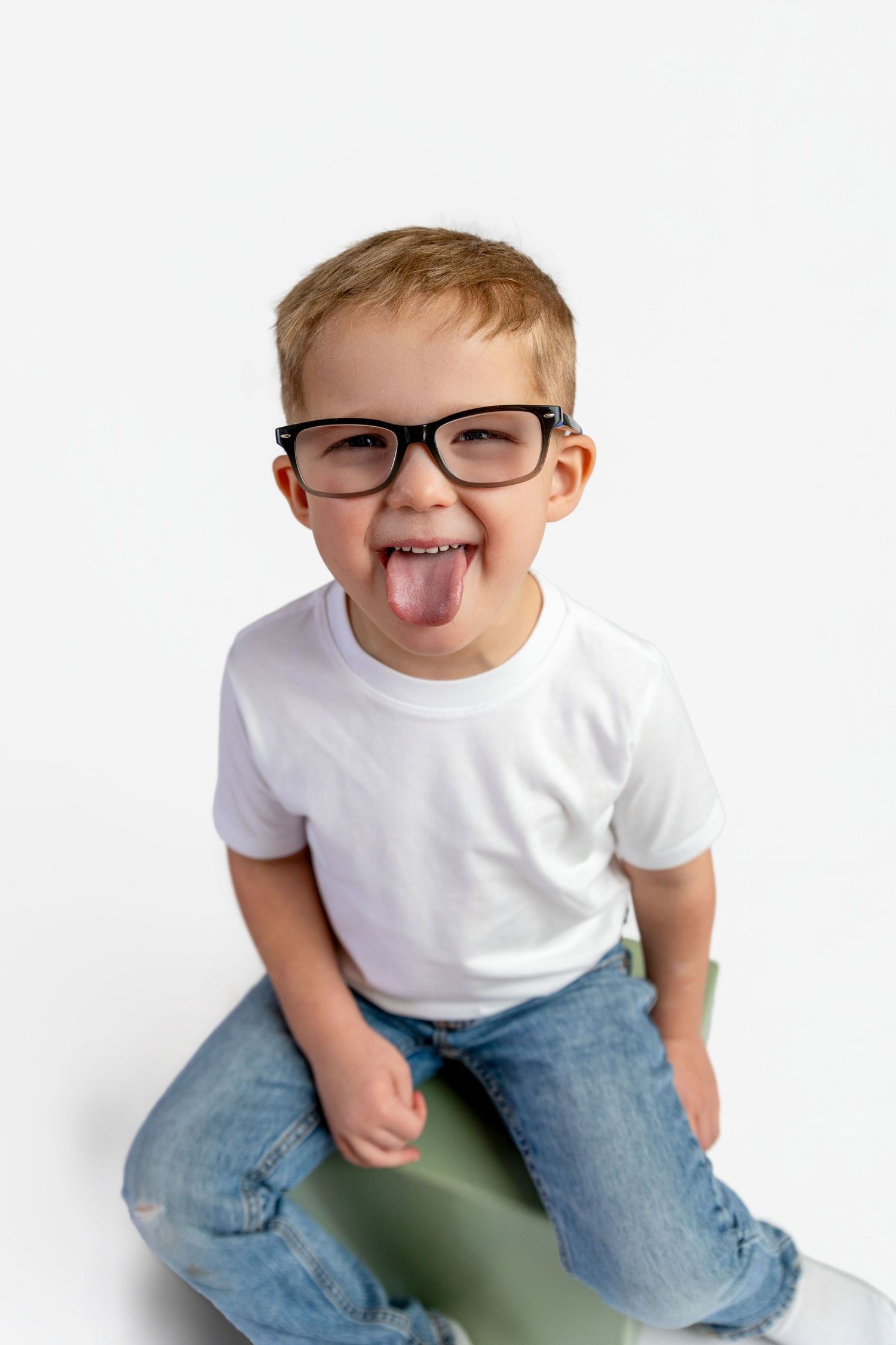 A playful young boy in glasses and white t-shirt sticking his tongue out while sitting against a white background.