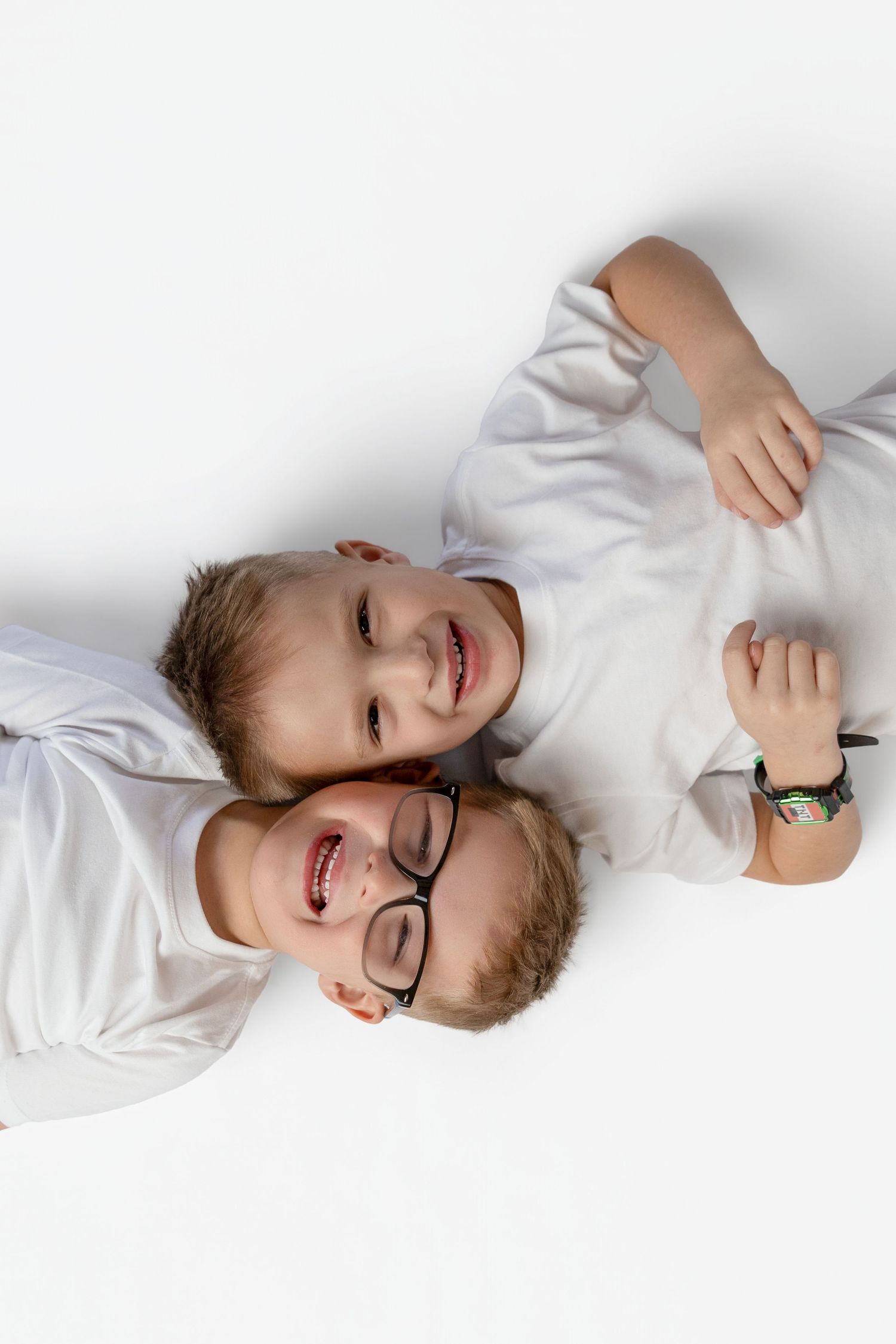 Two smiling boys in white shirts lying on a white floor, one wearing black glasses and a colorful bracelet.
