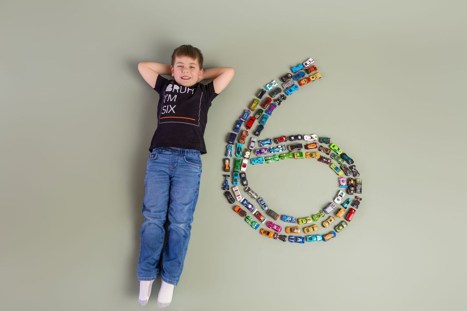 A boy lying next to a number 6 made of toy cars, celebrating his 6th birthday.