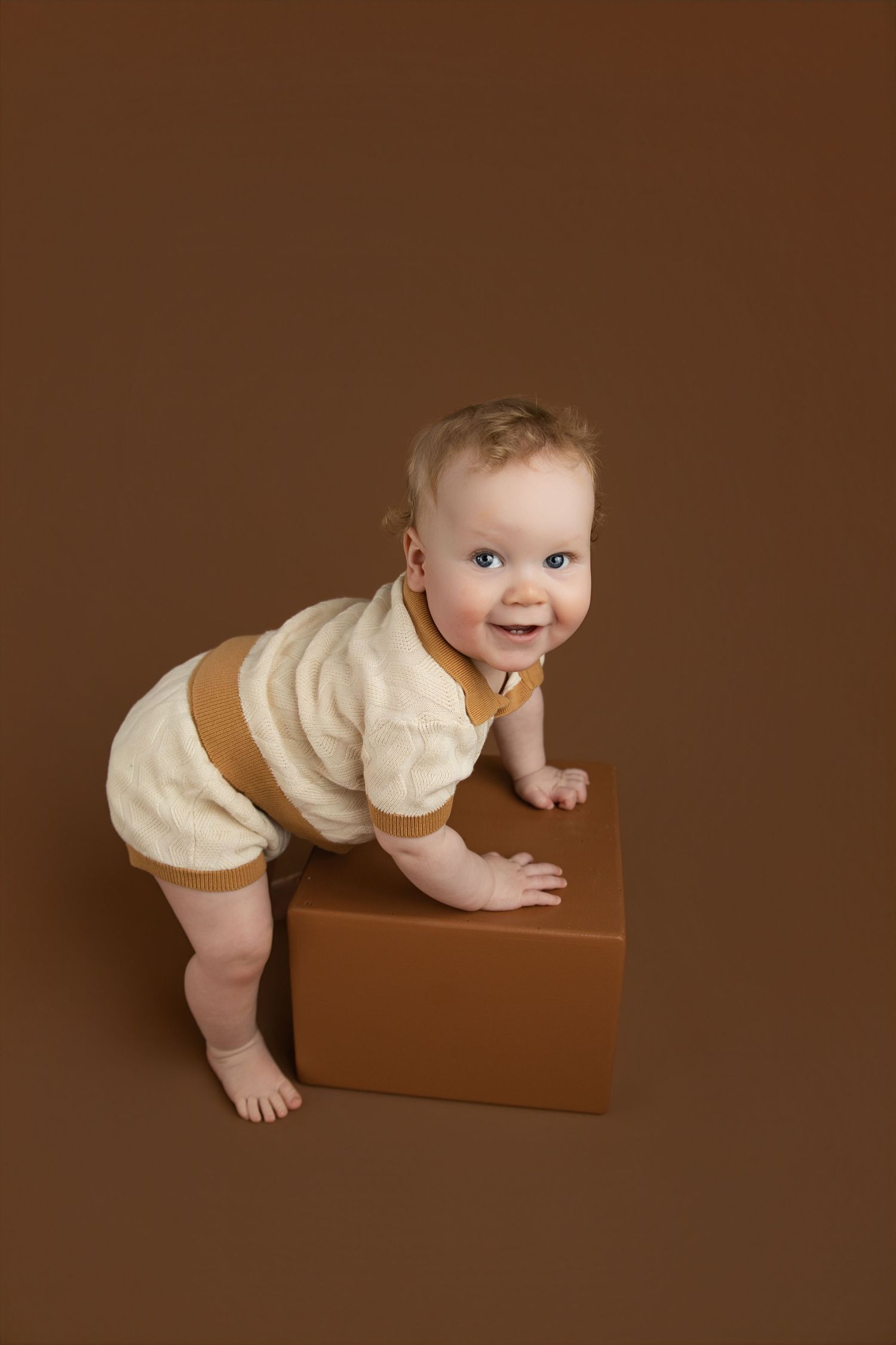 Smiling baby in cream and brown romper leaning on a brown cube against a matching brown background.