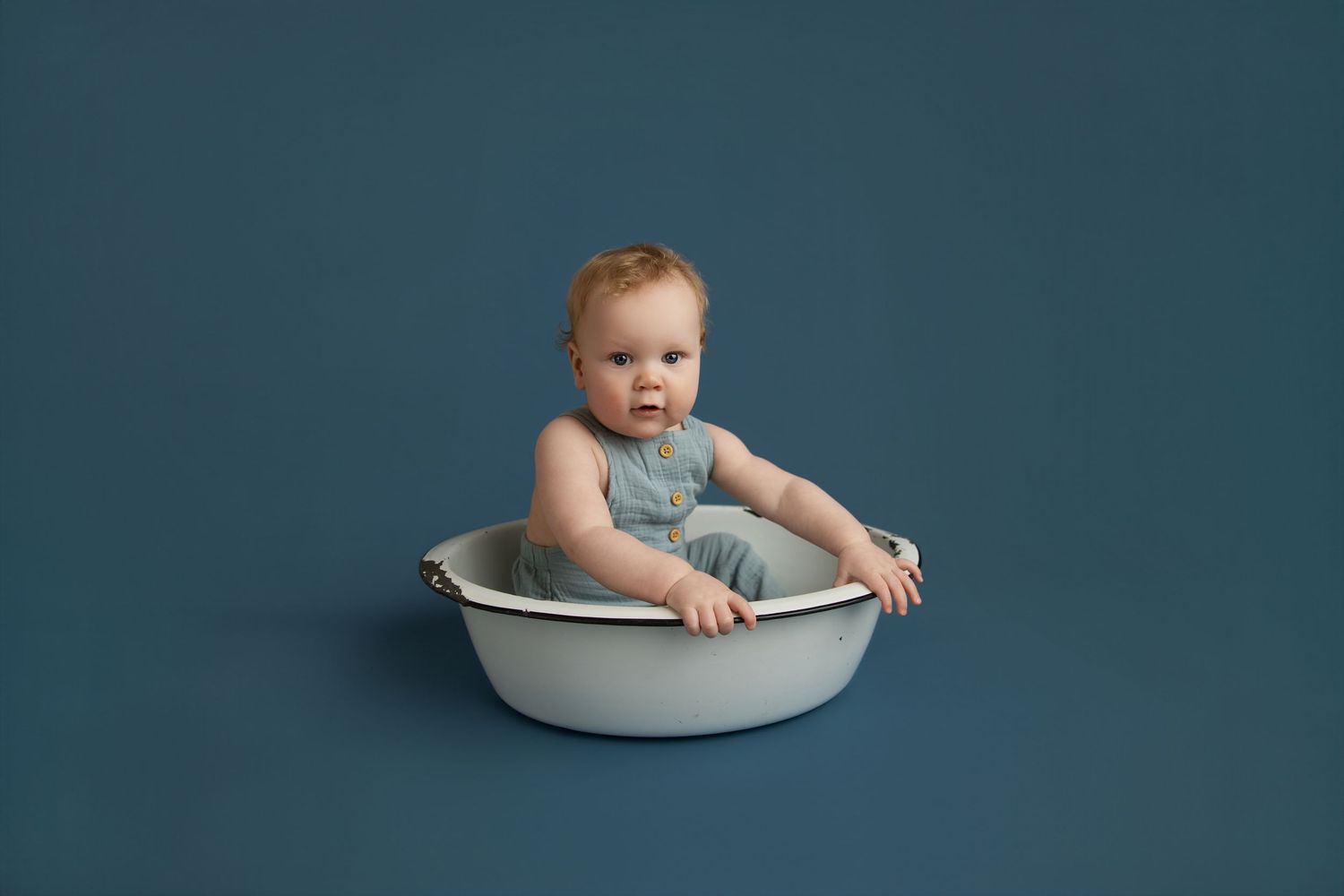 A baby boy in a blue romper sits in a vintage white enamel basin against a deep blue background.