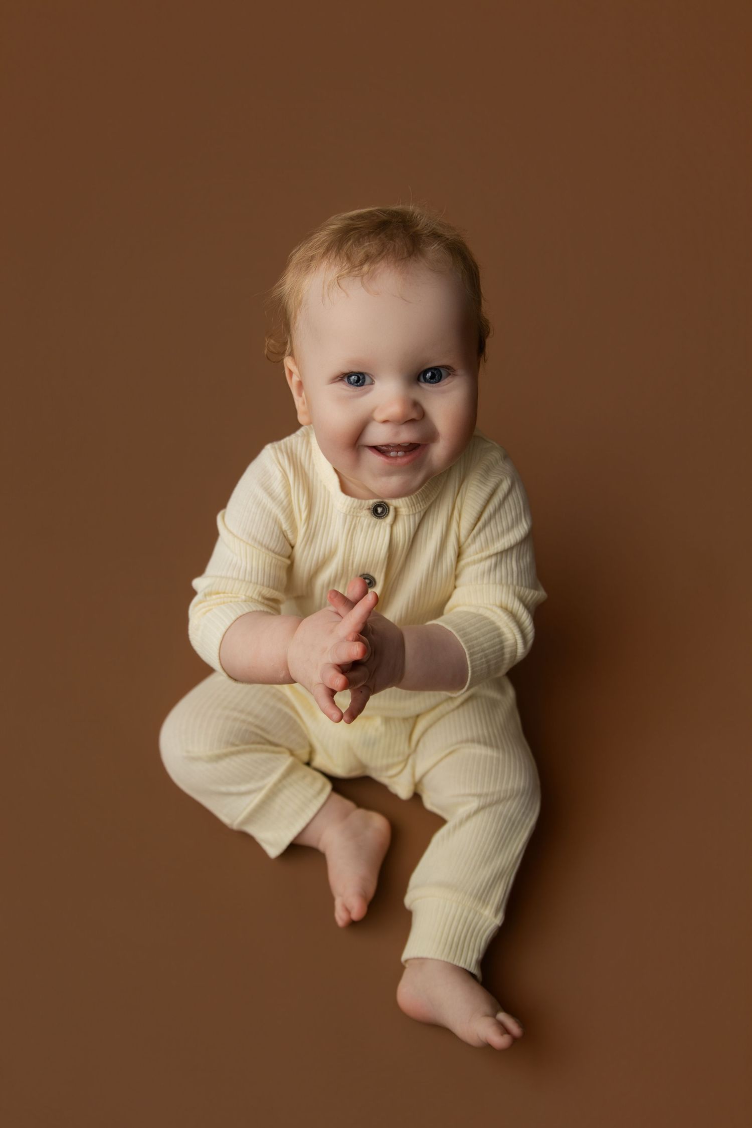 Smiling baby in cream ribbed romper sitting on brown background with blue eyes and reddish hair.