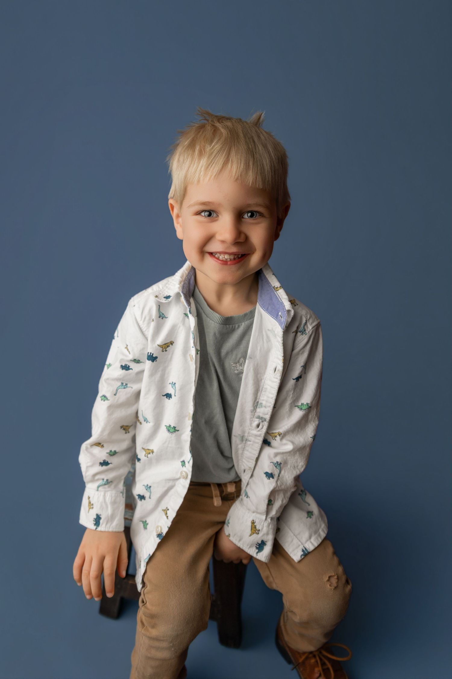 Smiling blonde boy in patterned white shirt and khaki pants posing against blue background.