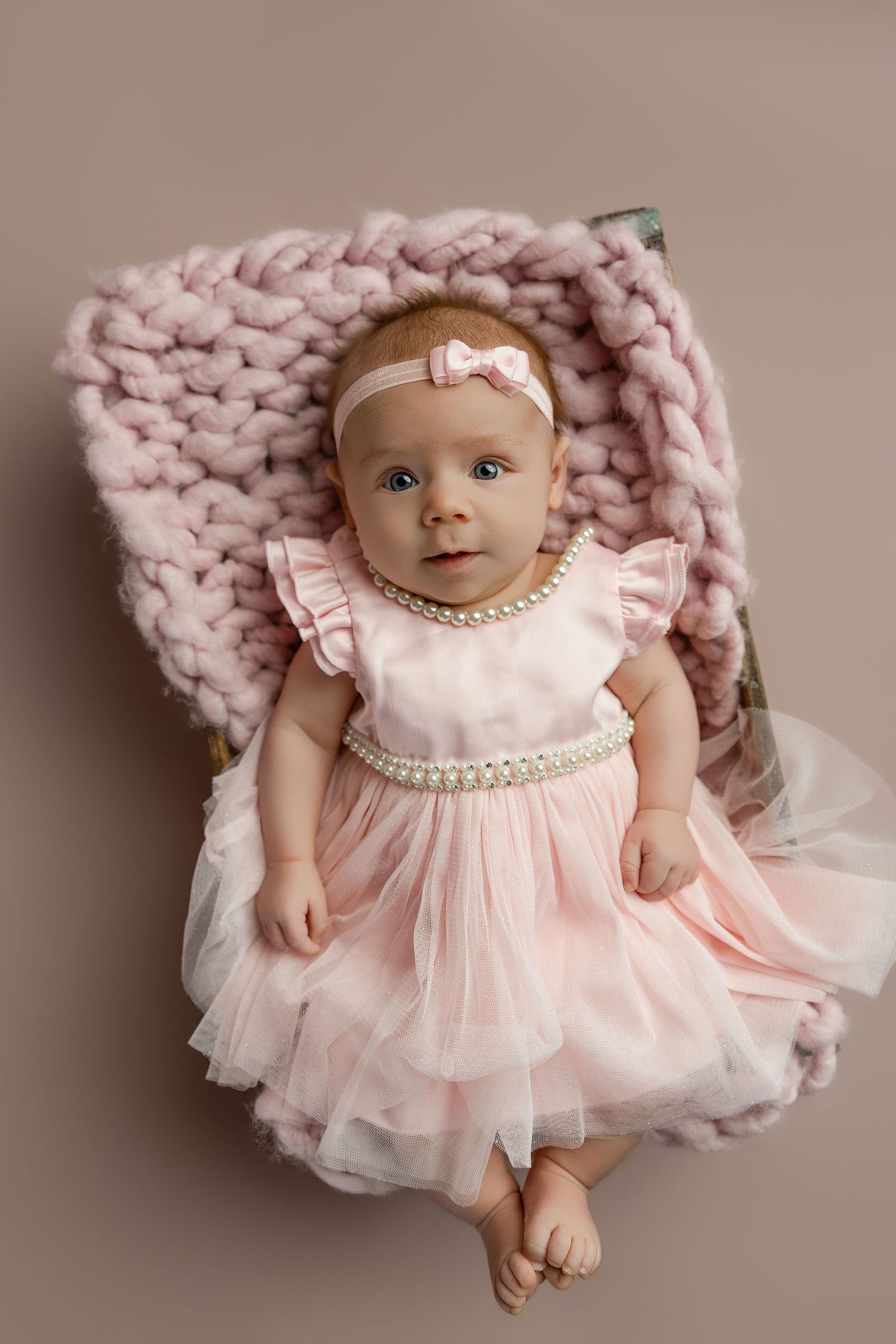 A baby girl in a pink tulle dress with pearl necklace lies on a chunky knit blanket for a professional portrait.