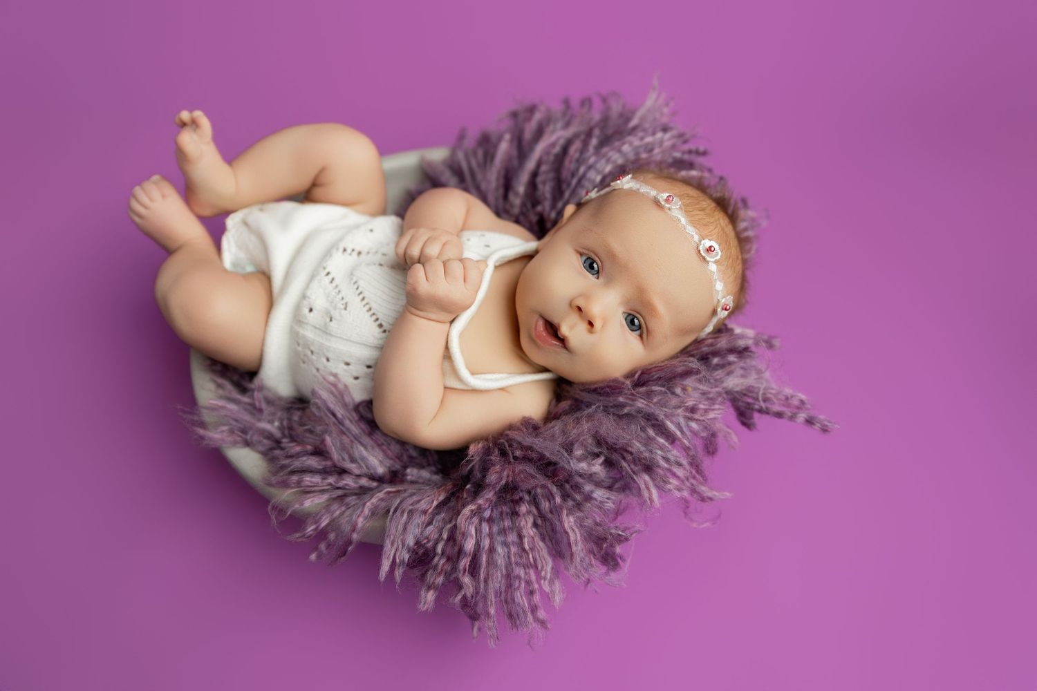 Smiling baby in white romper lying in purple fluffy bowl on lavender background.