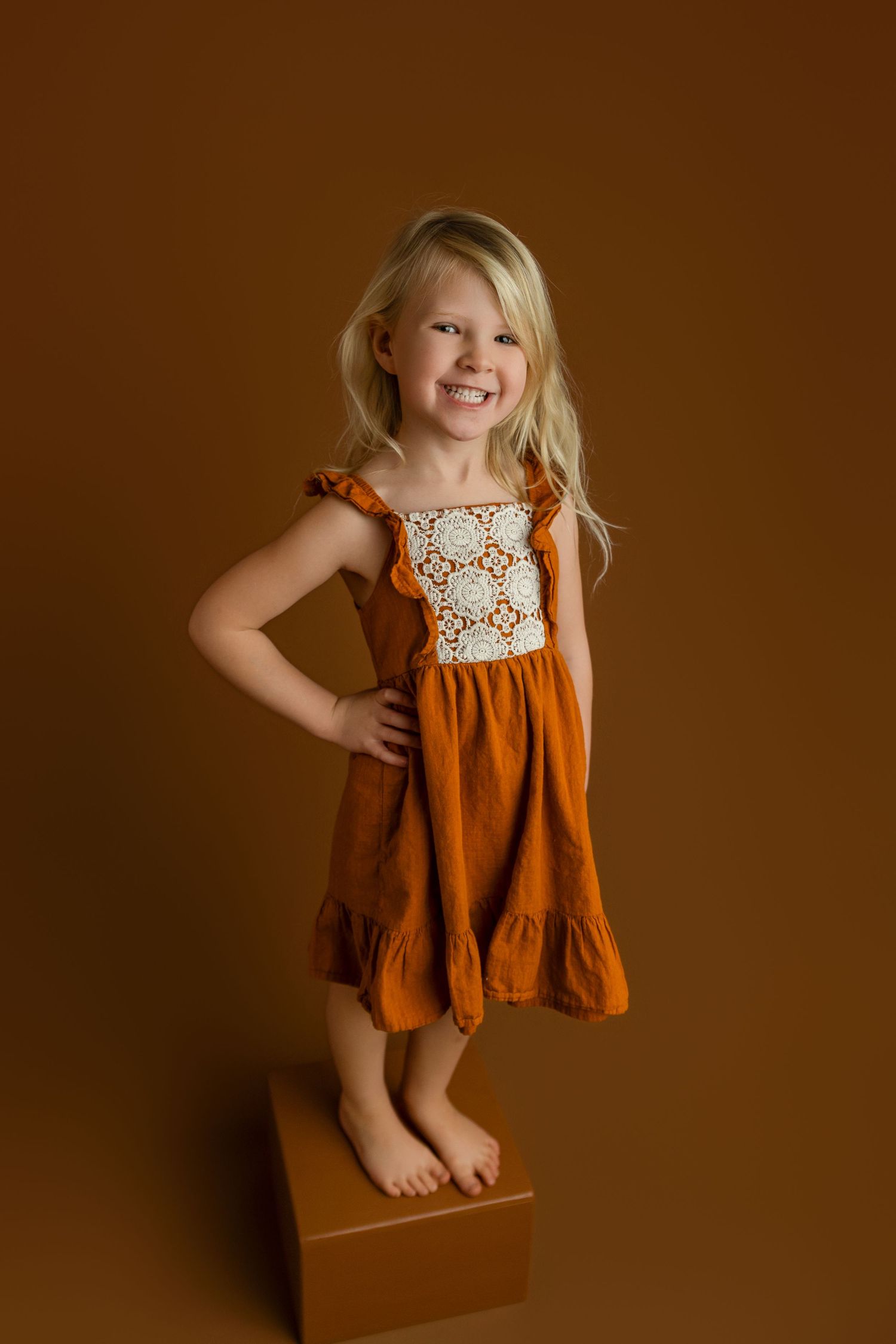 A smiling blonde girl in an orange rust dress with lace detail stands barefoot on a wooden box against a brown background.