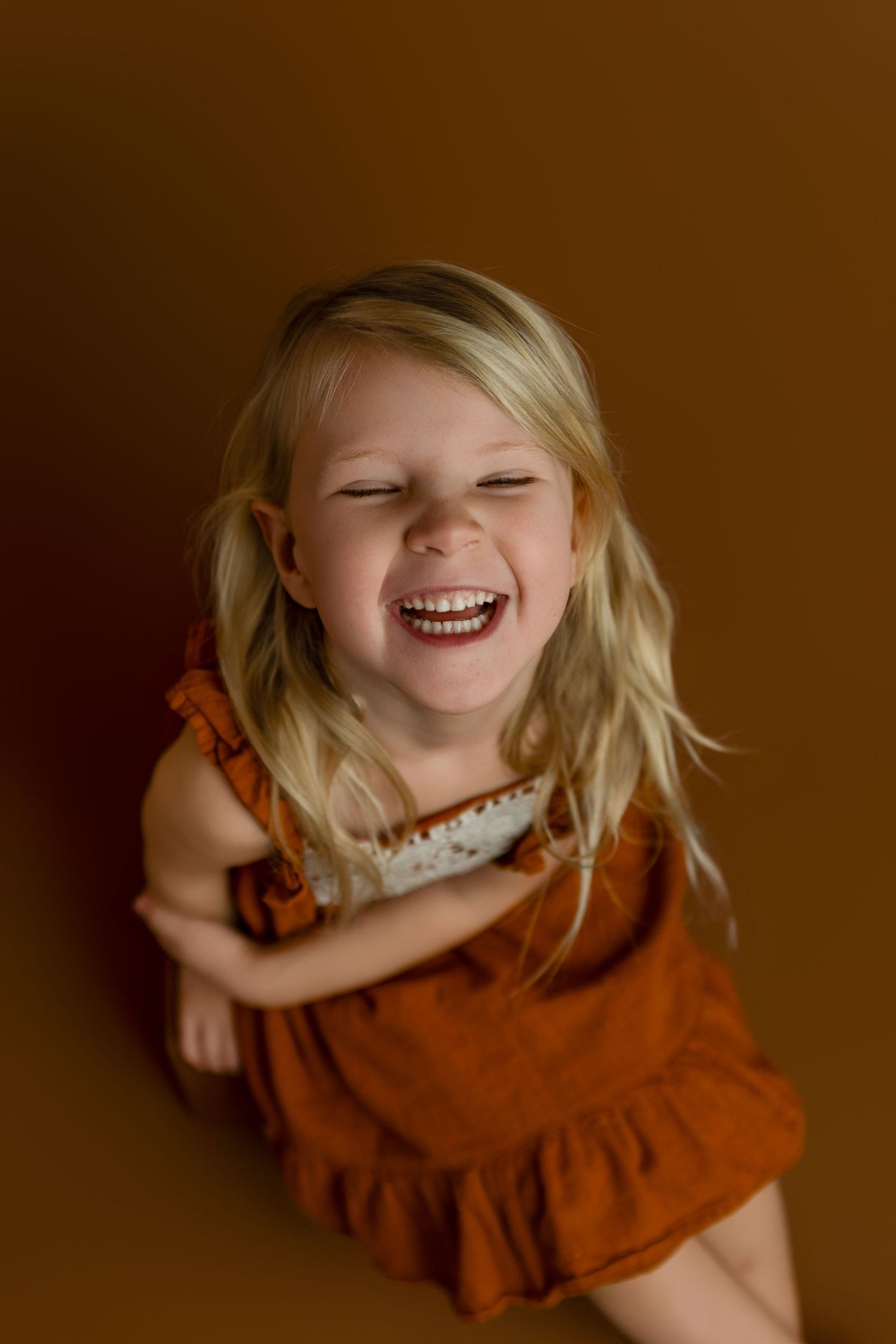 A joyful blonde toddler girl in a rust orange dress laughs with eyes closed against a warm brown background.