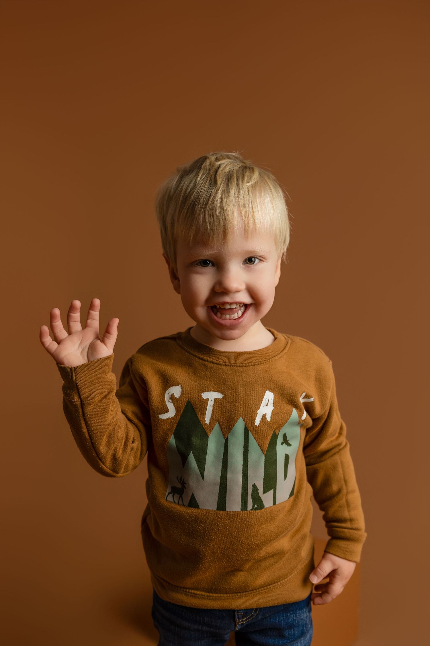 Happy toddler boy with blonde hair wearing a brown Stay Wild sweatshirt, waving and smiling at the camera.