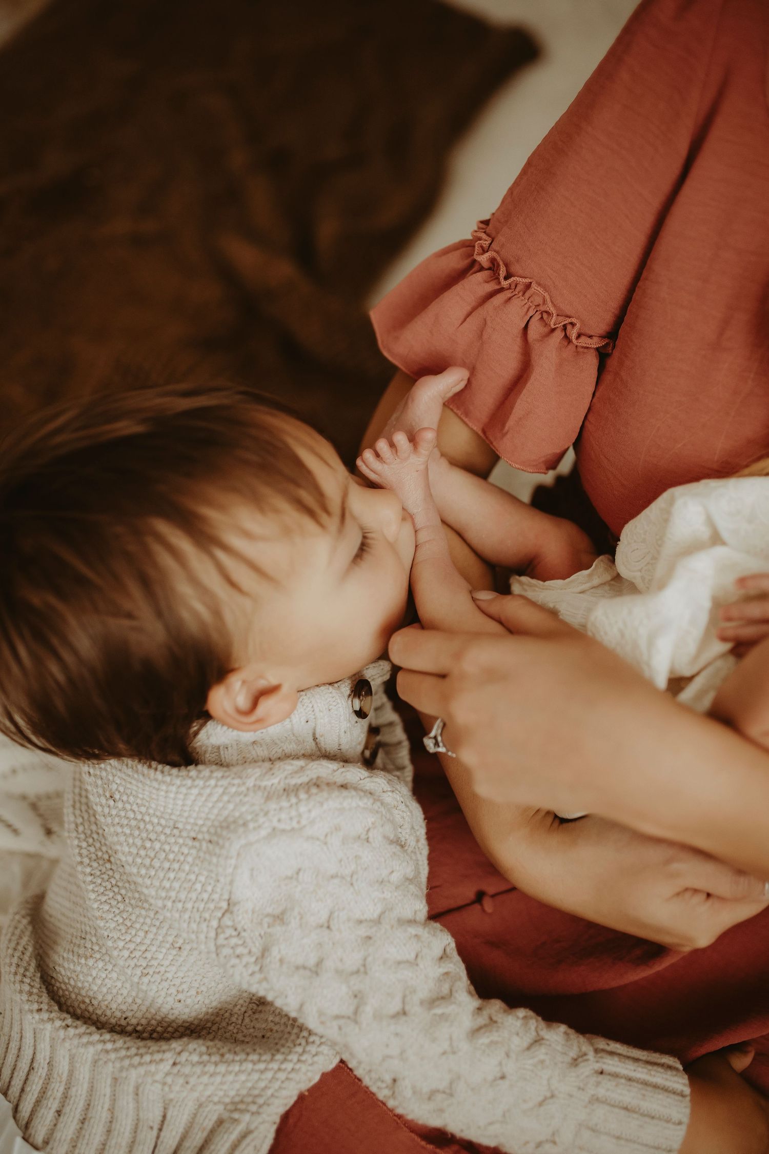 A tender breastfeeding moment captured in warm tones with a white knit blanket and rust-colored clothing.