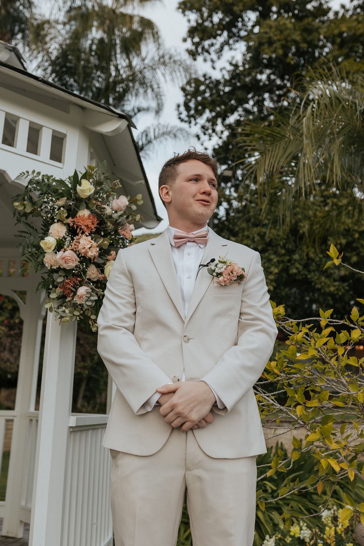 A groom in a cream colored suit and pink bow tie stands in front of a floral arch at an outdoor wedding venue waiting for his bride.