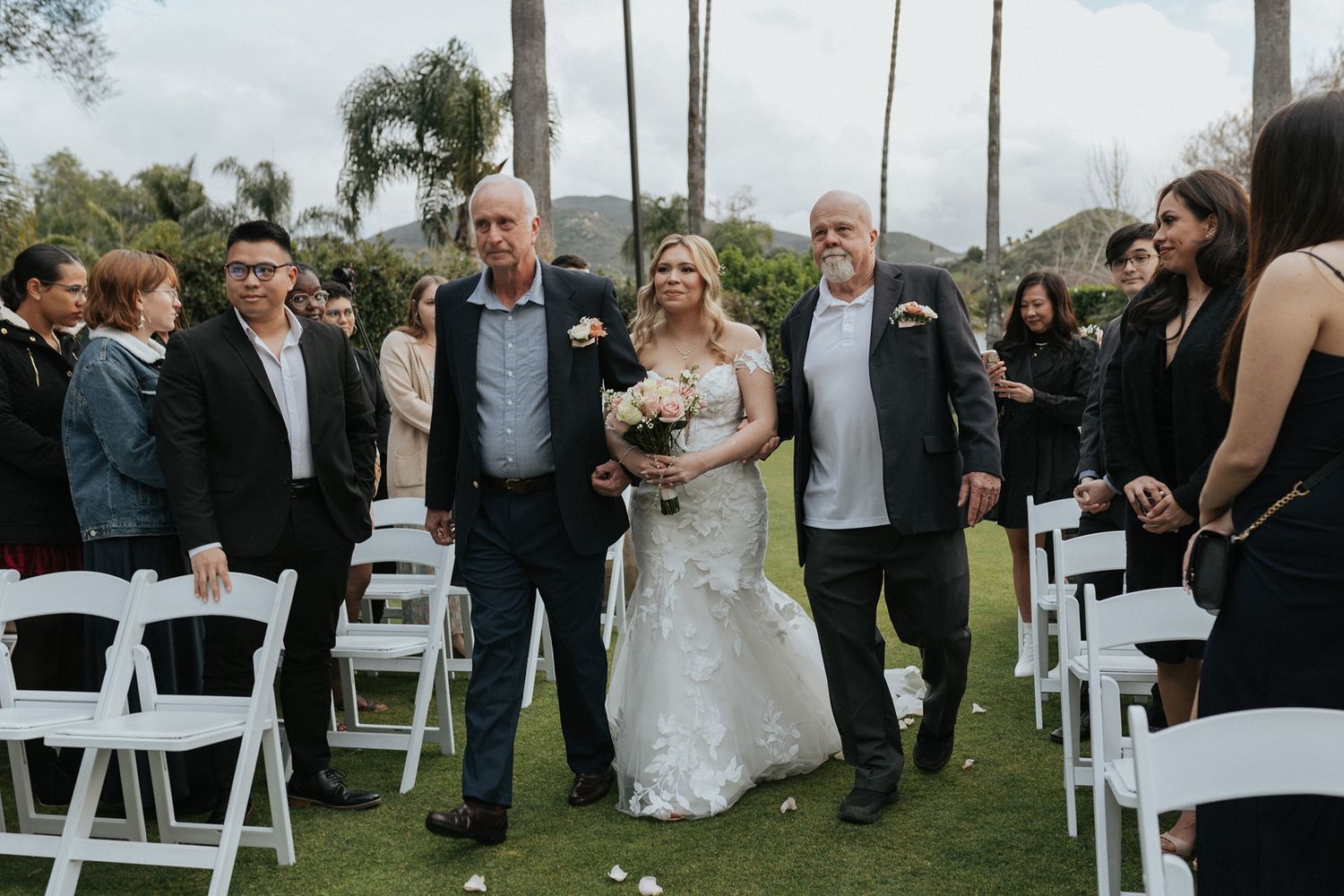 A bride walks down the aisle at a ceremony on a lush green lawn with white chairs and palm trees in the background.