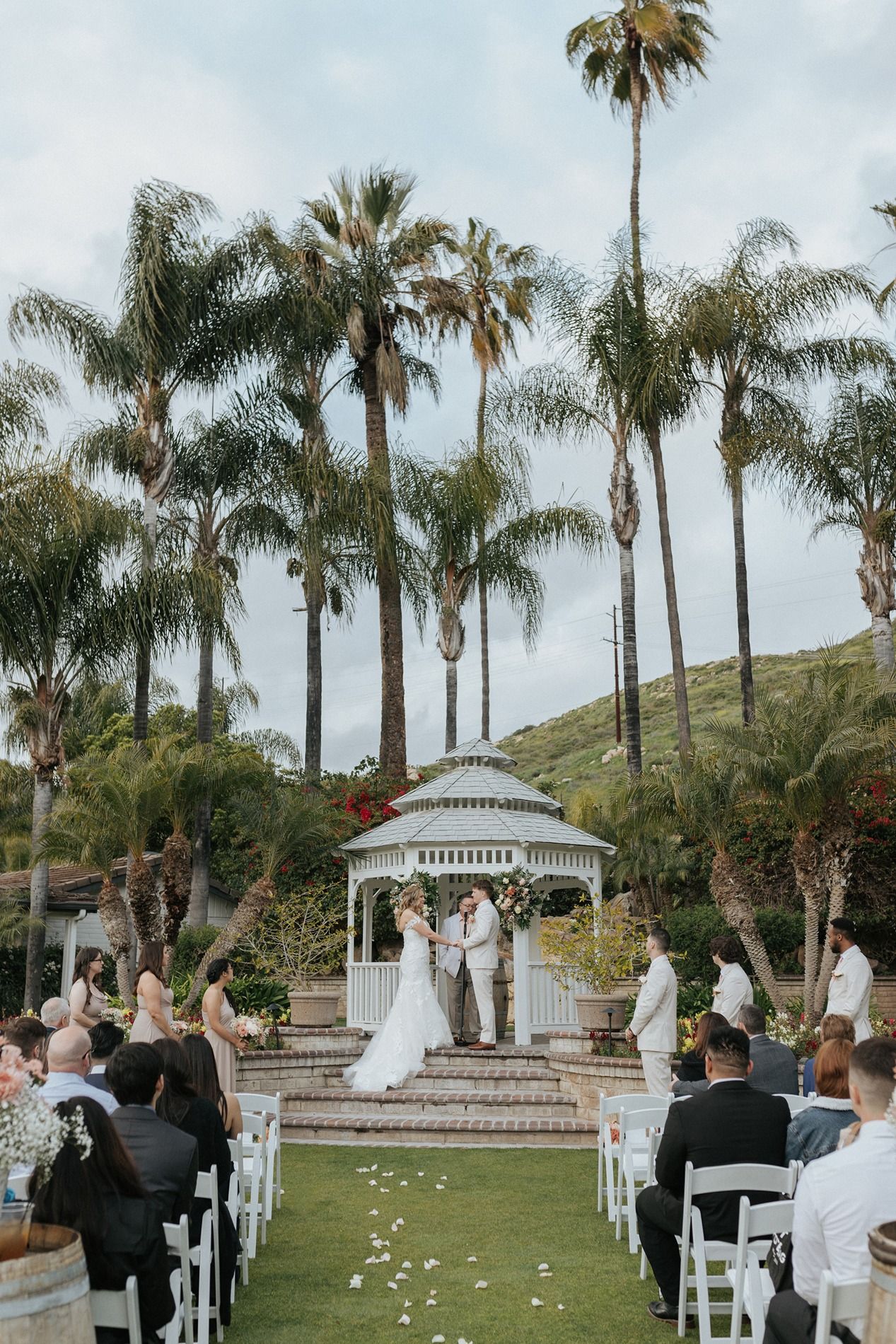 Outdoor wedding ceremony setup with white chairs and gazebo surrounded by palm trees on a grassy hillside.