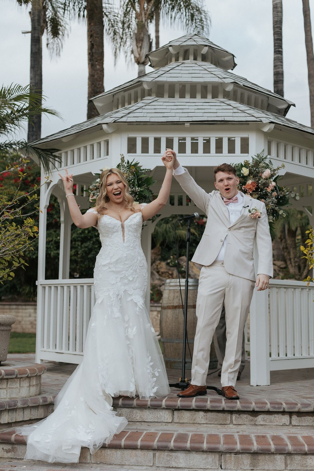 A couple celebrates their wedding day with joyful poses in front of a white gazebo at an outdoor venue.