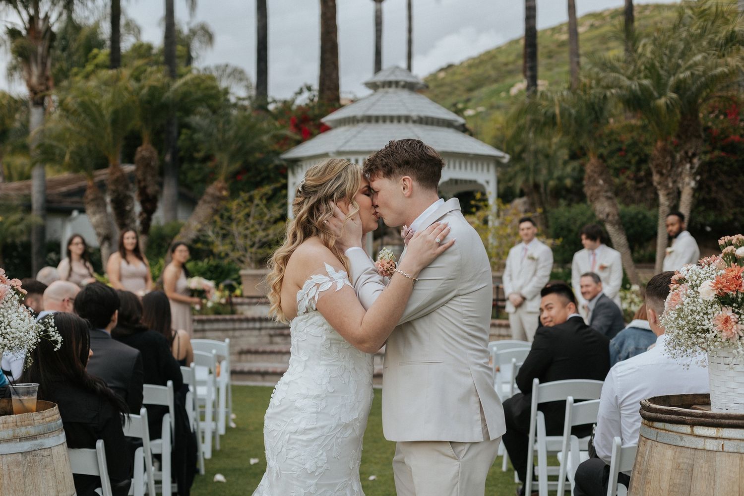 A couple shares their first kiss at an outdoor wedding ceremony with guests seated in white chairs and a gazebo backdrop.