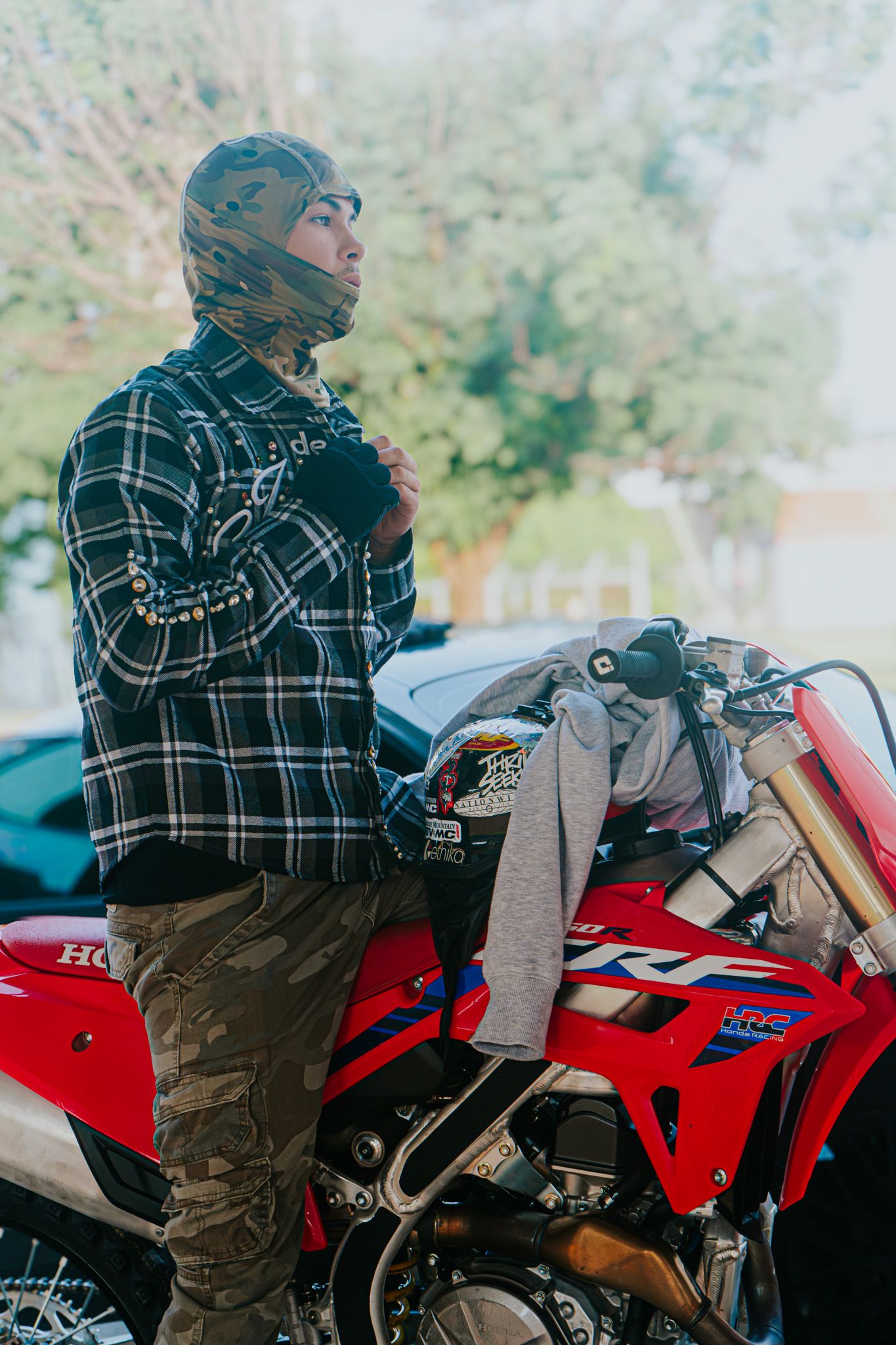 Motorcyclist in plaid shirt and protective gear seated on red dirt bike.