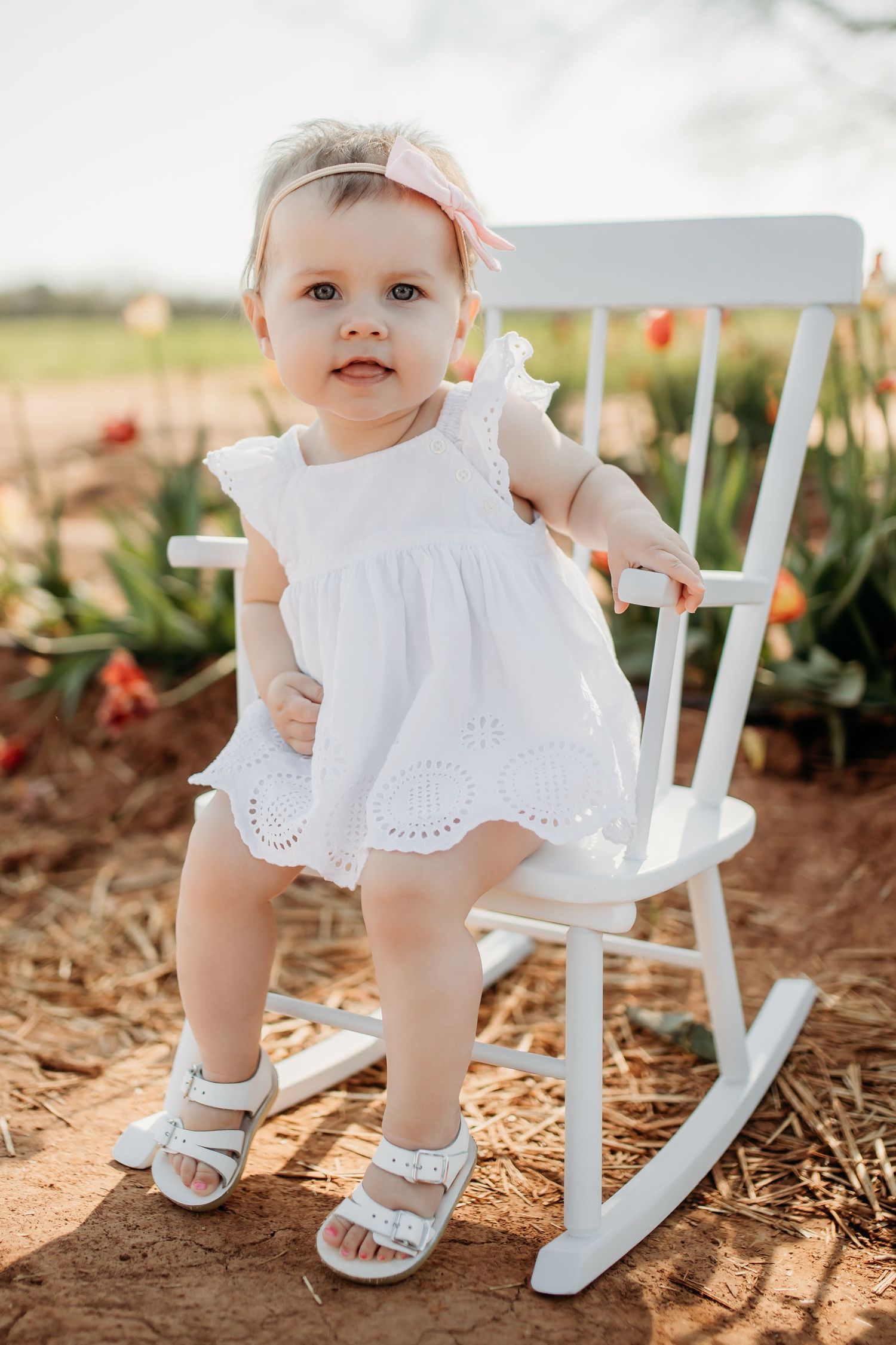 Close-up views of tiny feet in white shoes next to a white rocking chair on dirt ground in sunlight.
