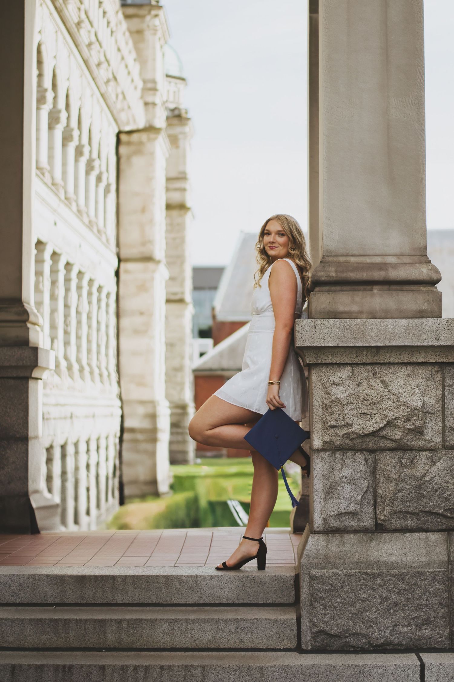 A woman in a white top and blue skirt sits gracefully on stone steps between classical architectural columns.