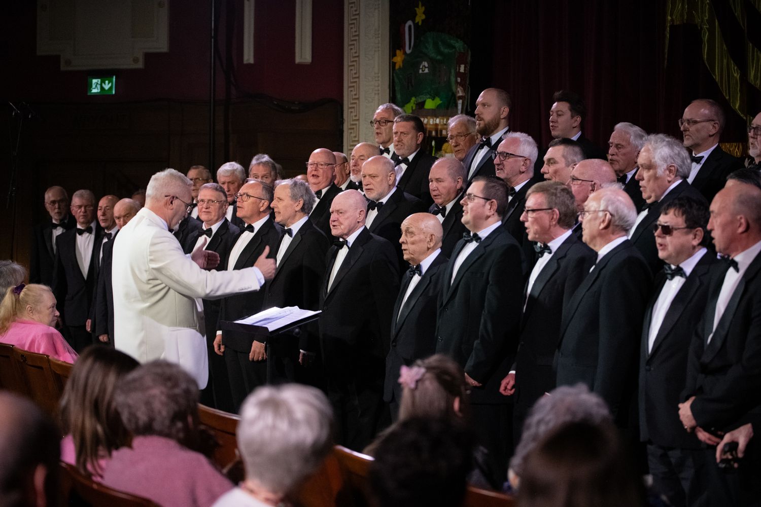 Male choir in formal attire performing on stage with conductor in white jacket. Event photography by K Lewis Photography.