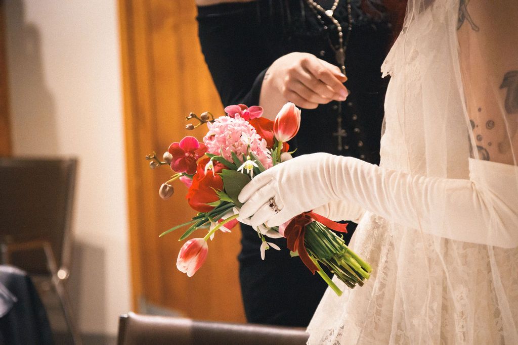 Bride in white lace dress and gloves holding vibrant red and pink tulip bouquet at wedding ceremony.
