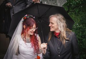 Bride with red hair and floral crown shares umbrella with partner in black suit on rainy wedding day.