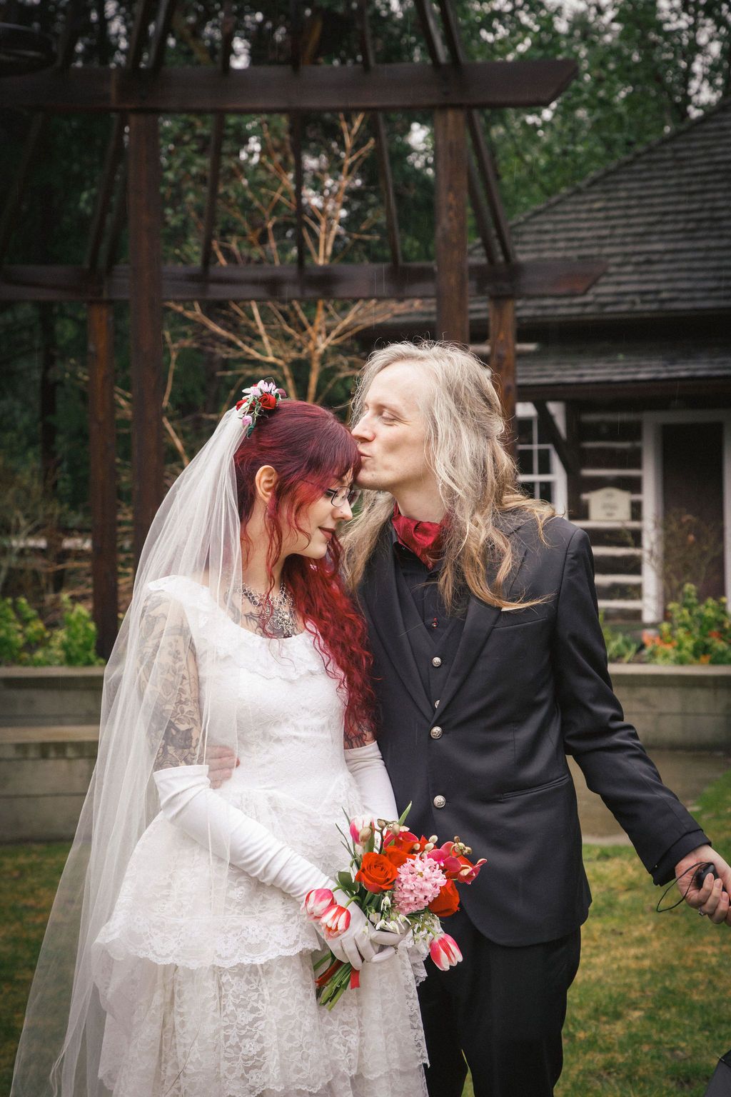 Groom kisses bride with red hair holding pink tulip bouquet outdoors at rustic garden wedding venue.