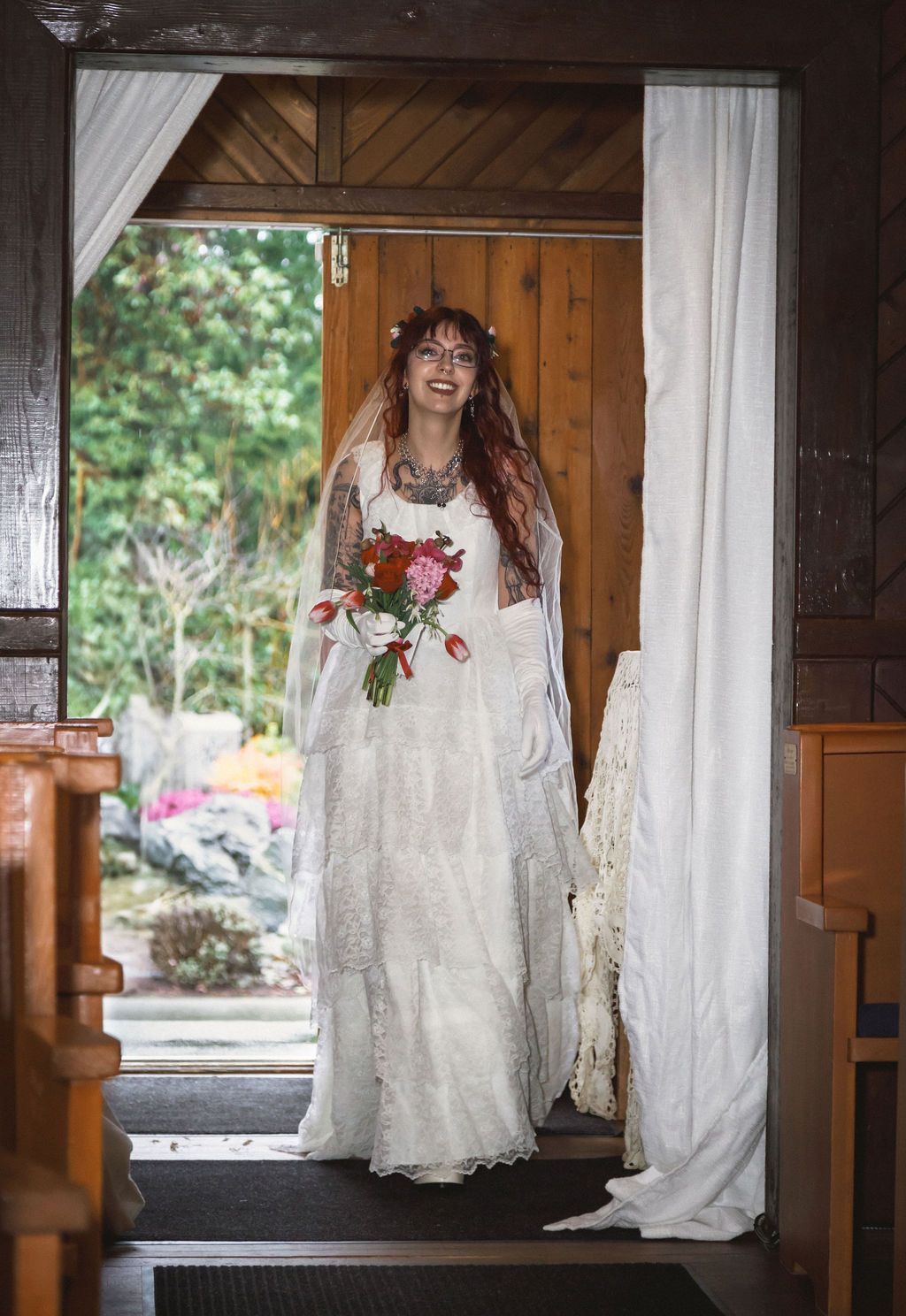 Tattooed bride in white lace gown and veil standing in chapel doorway holding red and pink bouquet.