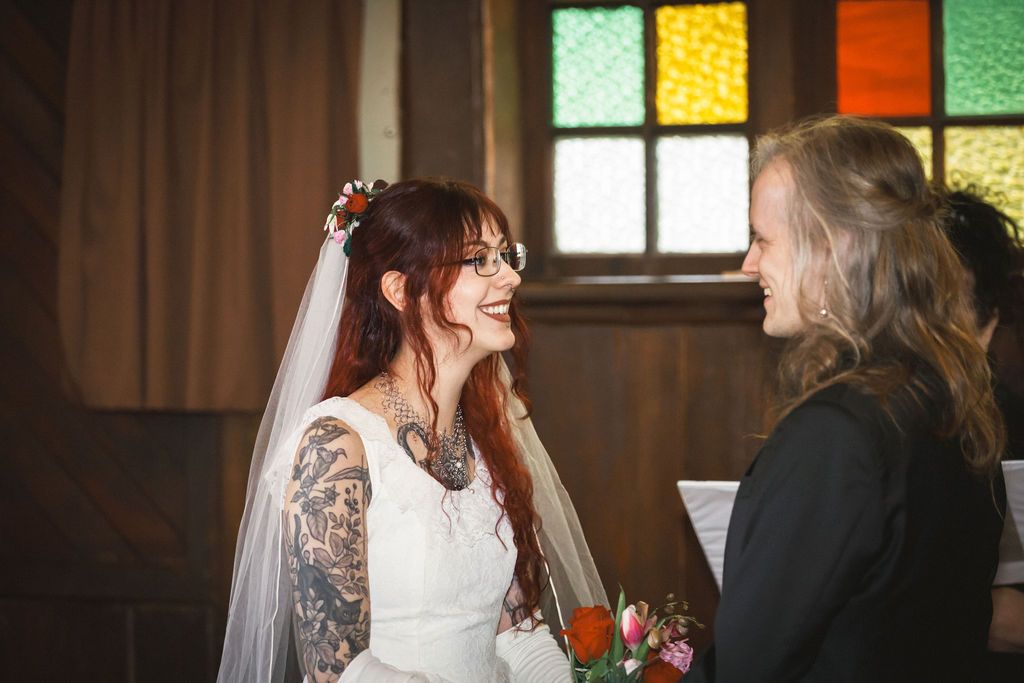 Tattooed bride in white veil laughing joyfully with officiant near colorful stained glass windows.