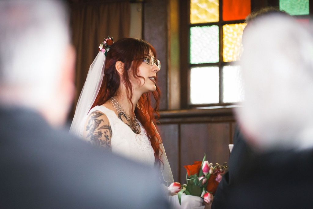 Tattooed bride in white veil smiling during wedding ceremony with colorful stained glass in background.