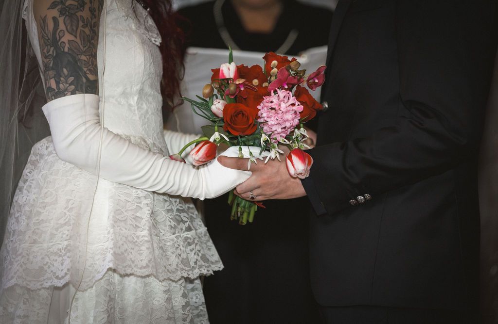 Bride and groom holding hands with vibrant red and pink floral bouquet during wedding ceremony.