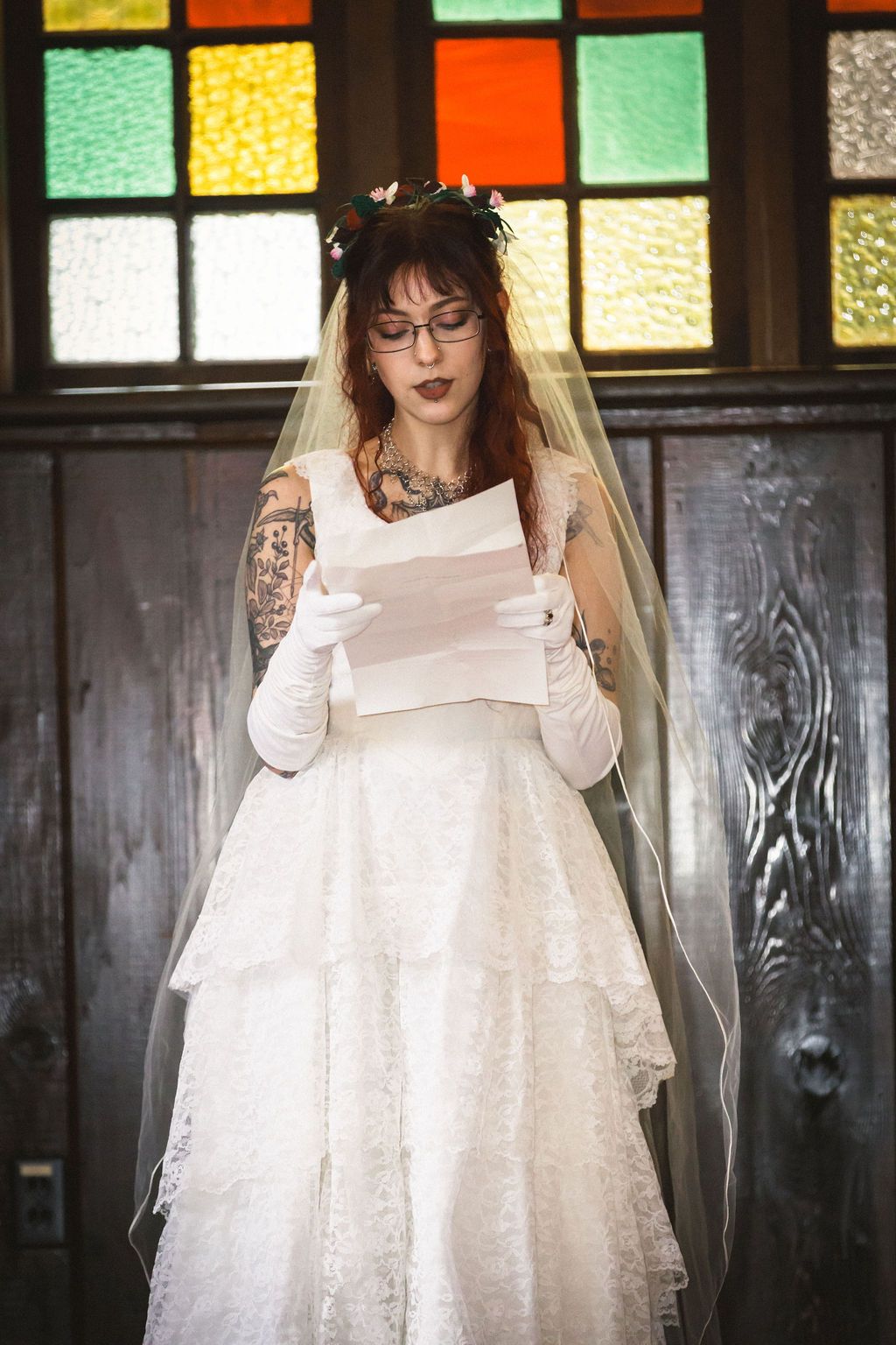 Tattooed bride in white lace gown and veil reads vows before colorful stained glass windows.