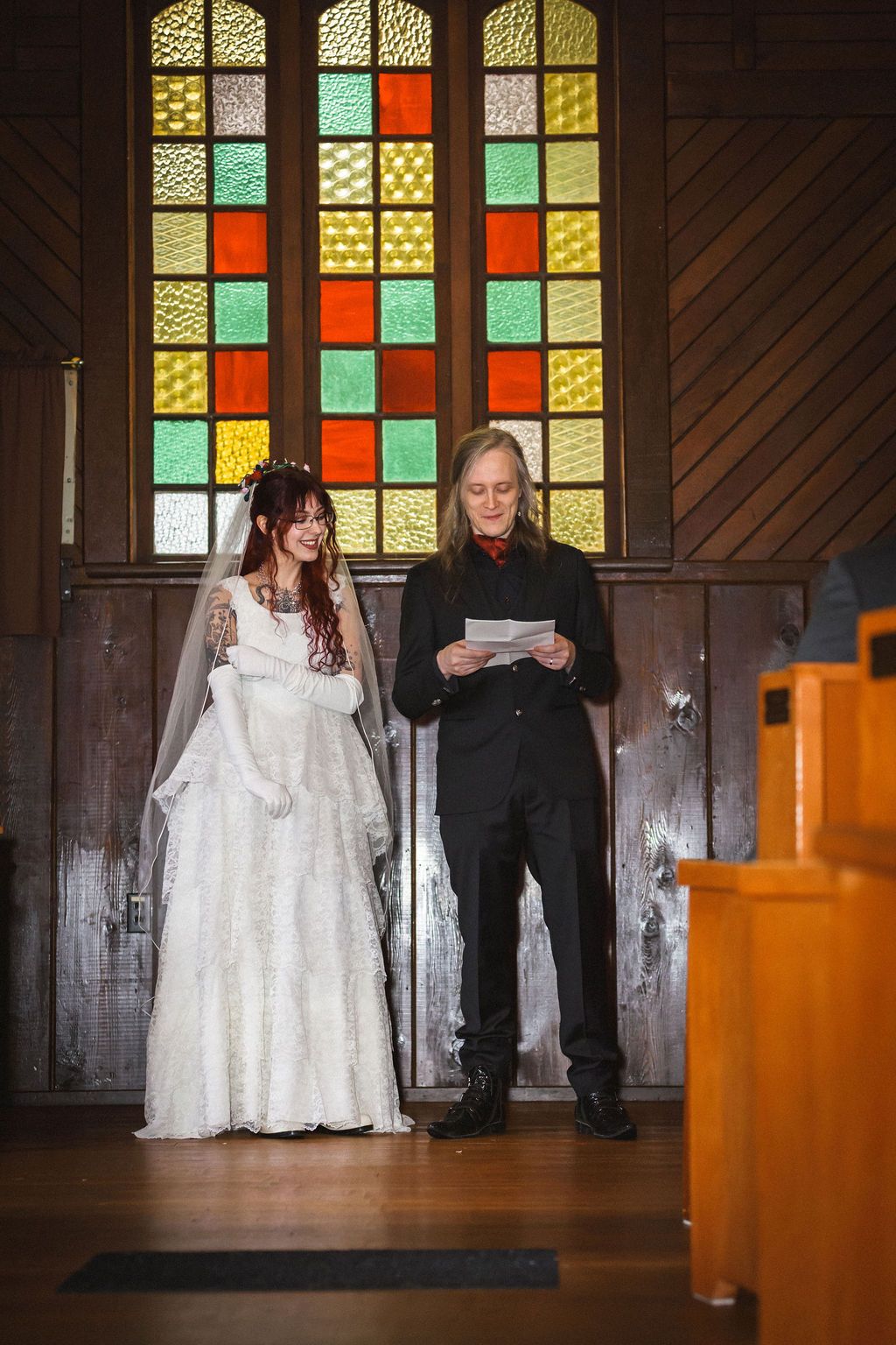 Smiling bride in lace gown and groom in black suit stand together at chapel altar.