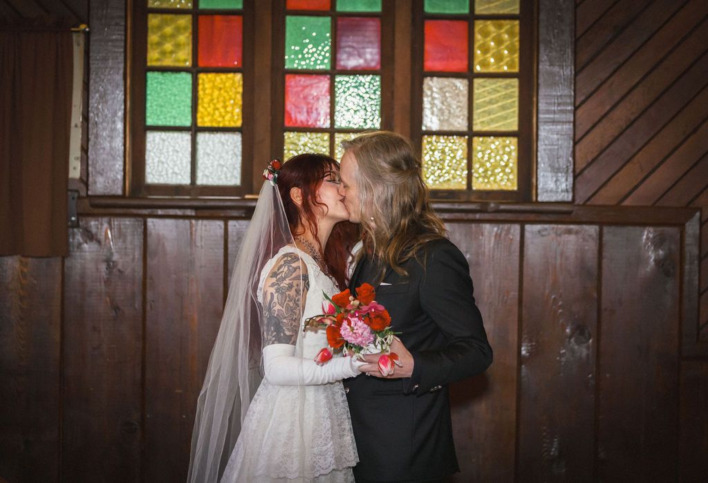 Bride and groom share a kiss in front of vibrant stained glass windows during wedding ceremony.