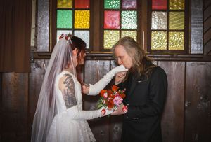 Groom kisses bride's hand while she holds colorful bouquet before stained glass windows.